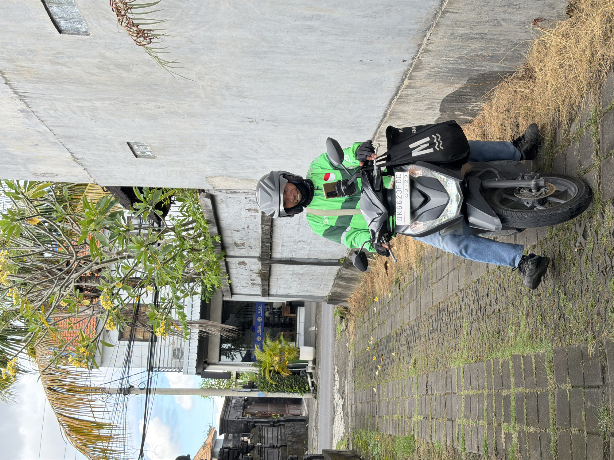 A man wearing a grey helmet and green jacket is sitting on a grey motorbike on a brick road in North Kuta, Indonesia. The license plate reads "DK 6623 FDC 03-29". He wears blue jeans and black shoes. A black bag with white markings is attached to the back of the bike. The street is lined with buildings, greenery, and utility poles.