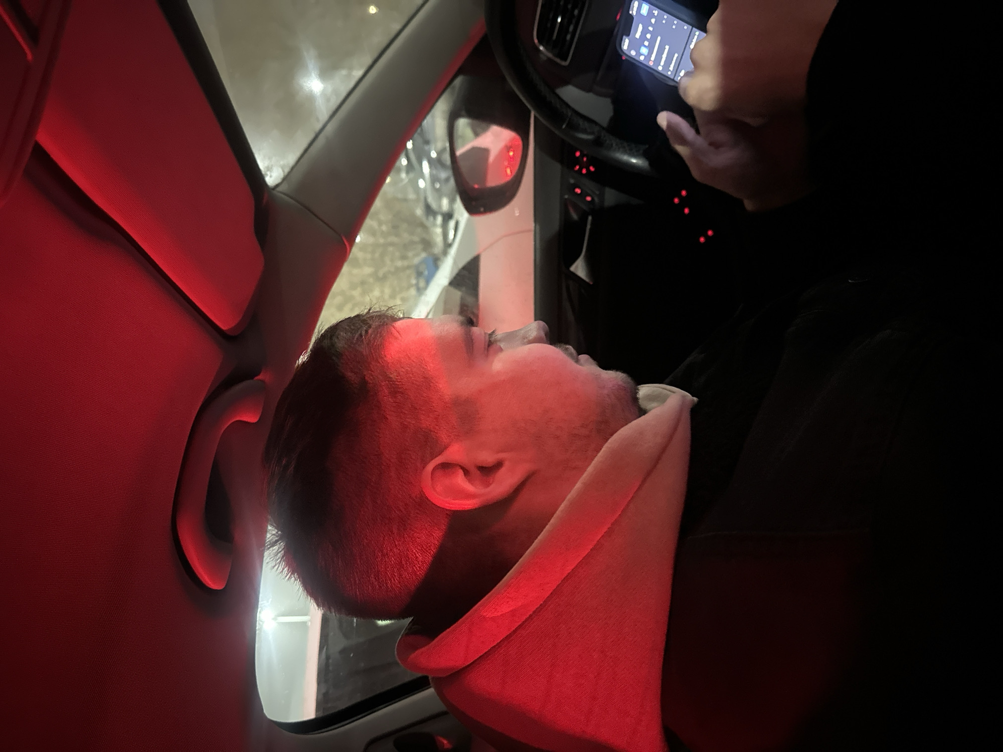 A side view of a man in a car, illuminated by red light. He appears to be either sleeping or unconscious, with his head resting against the headrest. A second person, only their hands visible, is holding and operating a smartphone in front of the steering wheel. The car is parked, with a side mirror visible reflecting other parked cars. The interior of the car is dark, except for the red lighting. The location is specified as Kazan, Russia.