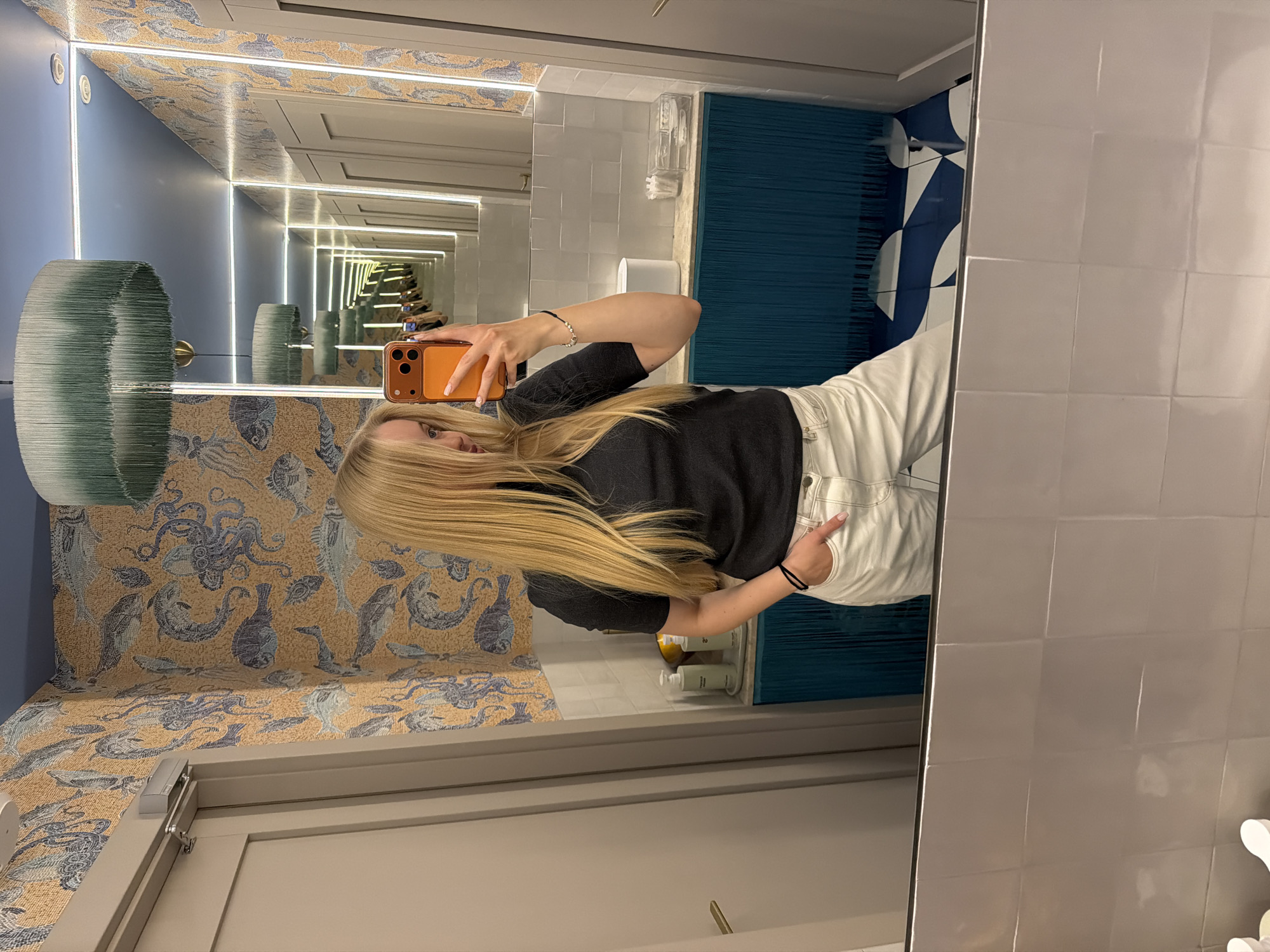A blonde woman is taking a selfie in a bathroom. She is wearing a black shirt and white pants. The bathroom has blue and white walls, and a wallpaper with sea creatures. There is also a mirror with a light around it. The woman is holding an orange smartphone in her right hand.