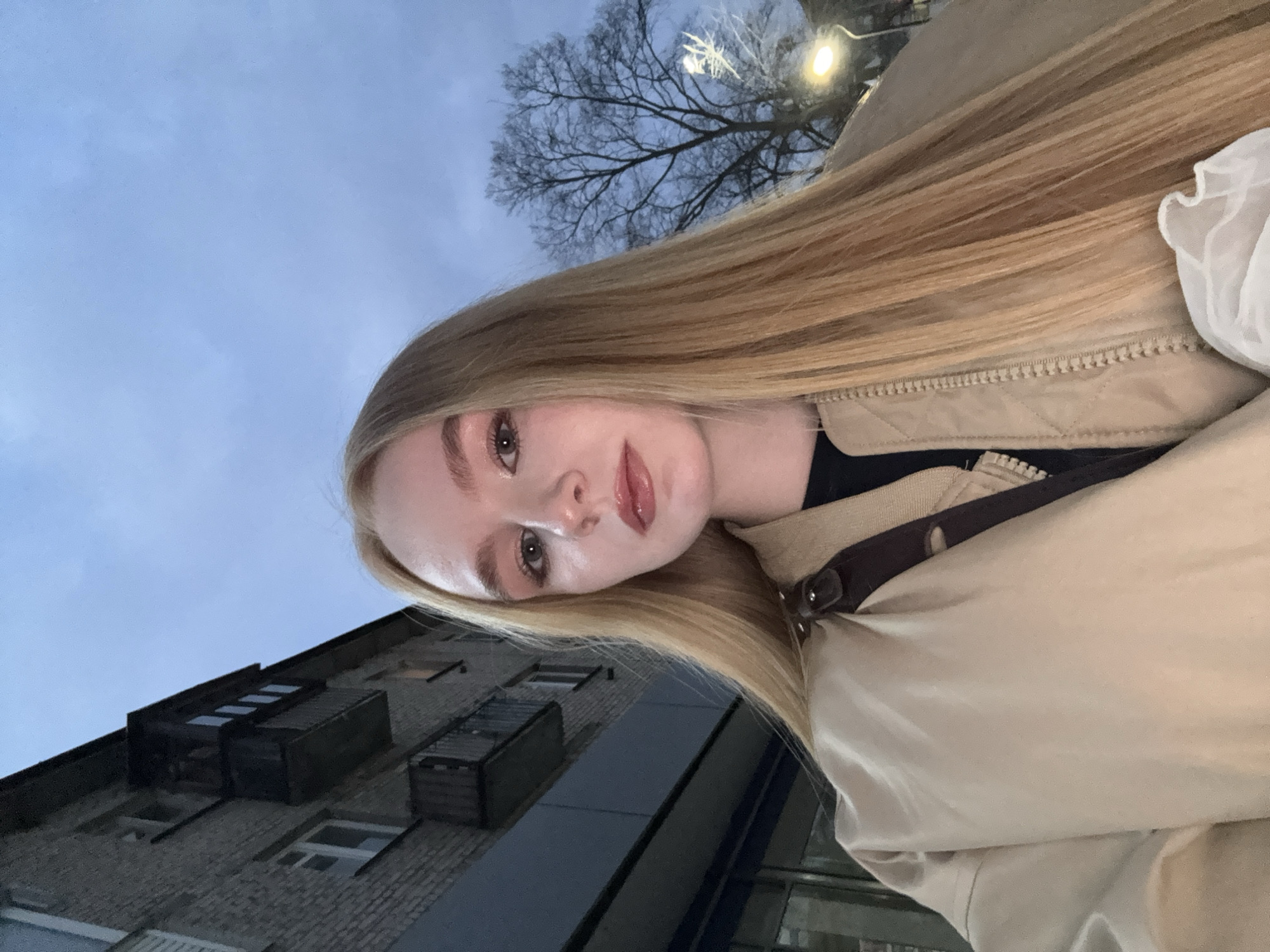 A selfie-style image is captured outdoors, featuring a young woman in an urban setting. The image is rotated 90 degrees clockwise, making the woman's head appear significantly tilted to the left.
The central subject is a woman with long, straight, blonde hair that cascades down the right side of the frame, covering part of her shoulder and extending towards the bottom. Her face is in the left-center of the image, angled slightly upwards towards the camera. She has light skin, light-colored eyes (possibly hazel or light brown), and her eyebrows are well-defined. She is wearing light makeup, including eyeliner and a glossy, pinkish lip color. Her expression is neutral to slightly serene, with her lips slightly parted.
She is wearing a light-colored (beige or tan) jacket, which has a visible zipper on the front, characteristic of a quilted or padded design. A dark strap, possibly from a shoulder bag or backpack, is visible across her chest. A glimpse of a white, ruffled fabric, possibly from a top or scarf, can be seen near her right shoulder in the upper right corner of the frame.
The background suggests a late evening or early night scene, indicated by the dim, overcast light blue-grey sky that occupies the upper portion of the frame. Artificial lights, appearing as bright, circular glows, are visible in the upper right, suggesting streetlights or building lights are on. A bare, deciduous tree with intricate branches is silhouetted against the sky in the upper right quadrant, indicating a colder season like late autumn or winter.
In the lower left background, a building made of light brown or grey brick is visible. It features several dark, rectangular windows and what appear to be dark-colored architectural elements or mechanical units on its upper levels or roofline. The building's structure is somewhat obscured by the woman's hair and body. The overall impression is an outdoor selfie taken in a city environment during dusk or nighttime.