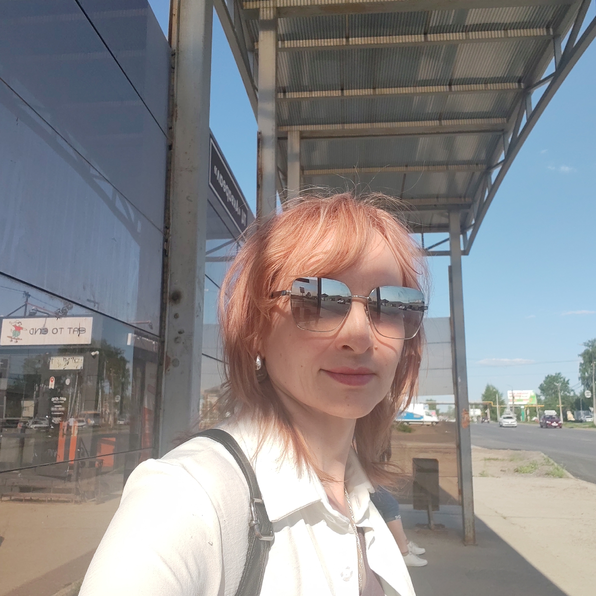 A woman stands at a bus stop in Chaikovsky, Russia. The bus stop structure is metal with a corrugated roof. A reflective building with storefront stickers is adjacent to the bus stop. The woman has reddish-blonde hair and wears sunglasses, a white collared shirt or jacket, and a small earring. She has a black backpack on her back. The sunglasses reflect the image of cars on a road. A road and sidewalk are visible beyond the bus stop. The sky is blue.