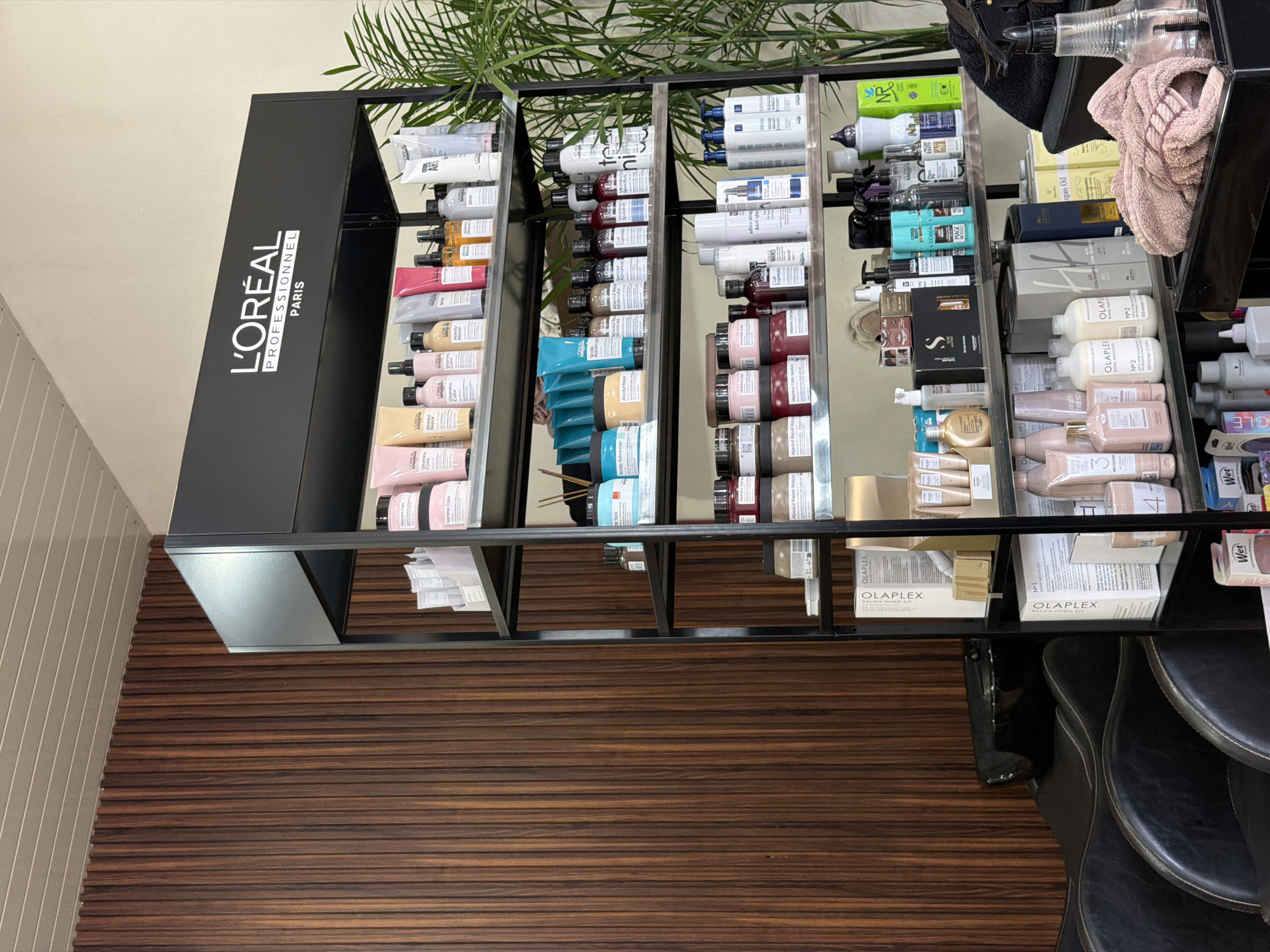 The image captures a beauty supply display, seemingly within a salon or retail setting. The prominent brand "L'Oréal Professionnel Paris" is vertically displayed on the black shelving unit. A variety of hair care products, including shampoos, conditioners, and styling aids from brands like L'Oréal, Olaplex, and others are neatly arranged on the shelves. A salon chair is visible at the bottom right of the frame. A few hair styling tools are placed on the shelf near the products. The floor is wooden, with a horizontal stripe pattern. Part of a potted plant appears in the upper portion of the image. The location is potentially in a retail store or a beauty salon in North Kuta, Indonesia, given the likely availability of these brand-name hair products in such establishments.