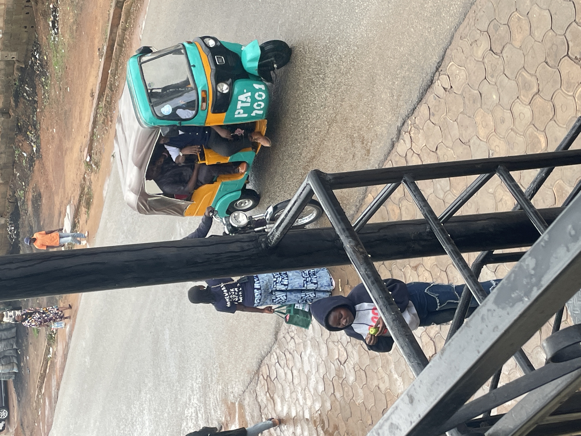The image depicts a street scene in Gura Top, Nigeria. In the foreground, a young person wearing a hooded sweatshirt and jeans is standing on a patterned walkway, holding a green object. A black metal railing is visible in the lower part of the frame. Further down the street, a woman in a colorful dress is walking, carrying a white bag. Behind her, another woman in a dark blue shirt and patterned pants is standing with two green bags. To the right, a turquoise and yellow auto-rickshaw is parked on the wet asphalt road, with three people inside. The auto-rickshaw has the registration number "PTA-1001" displayed on its side. An orange-shirted man is walking on the road in the background, and a motorcycle is parked to the left of the auto-rickshaw. The environment appears to be a typical urban street with some debris visible on the side of the road.