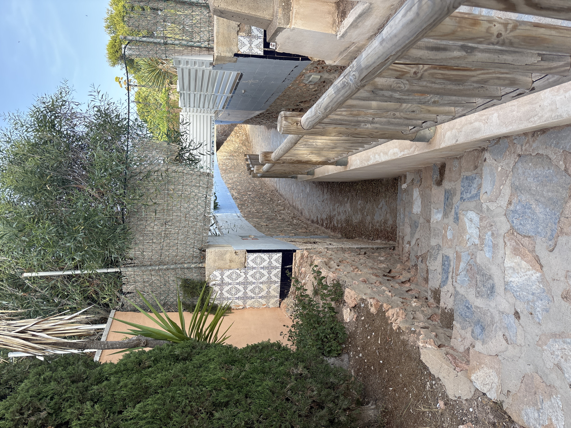 The image depicts a narrow pathway leading downwards, lined with a rustic wooden railing on the right. The pathway is paved with irregular stone slabs. The walls flanking the pathway are constructed from rough-hewn stones and feature sections of tilework. To the left, there is a dense green tree and a peach-colored wall with a yucca plant. The pathway appears to be in a somewhat rural or older urban setting, possibly within the municipality of Orihuela, Spain, given the architectural style and vegetation. The sky above is clear blue.