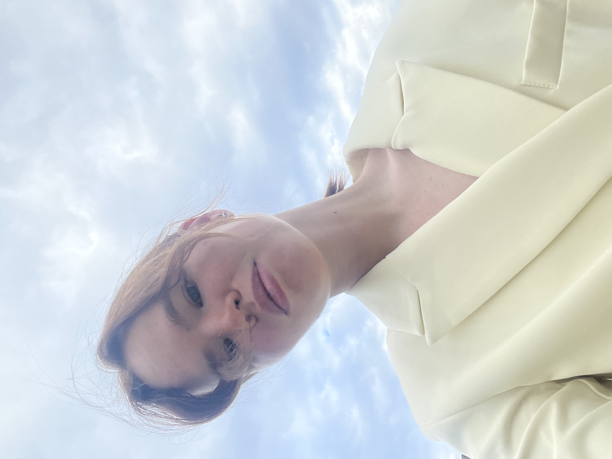 The image depicts a low-angle, eye-level selfie of a young adult female, captured outdoors in the city of Chaikovsky, Russia. The subject has light skin and reddish-brown hair, which appears slightly wind-blown, with some strands across her forehead and face. Her eyes are light-colored, and her lips have a natural, subtle coloring. A small, silver-colored hoop earring with a star-shaped charm is visible in her right ear. She is wearing a pale yellow or cream-colored blazer, visible from the mid-chest area upwards, featuring structured lapels and a pocket detail on the right side. The background consists entirely of a light blue sky with scattered white and grey clouds, suggesting an outdoor, daytime setting with soft, natural light. The overall scene is static, focusing on the individual against the sky.