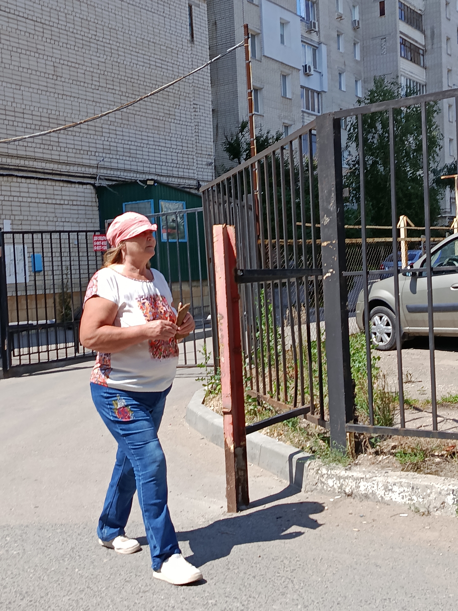 An adult woman walks down a paved path next to a black metal fence. She wears a light pink patterned headscarf, a white t-shirt with an abstract floral design on the chest and sleeves, blue jeans, and white athletic shoes. Her right hand is holding a small, flat, light brown object. Behind the fence, a silver car is parked. The background consists of a light gray brick building with multiple windows and a few trees. The scene appears to be in an urban setting in Saratov, Russia, given the architectural style and vegetation.