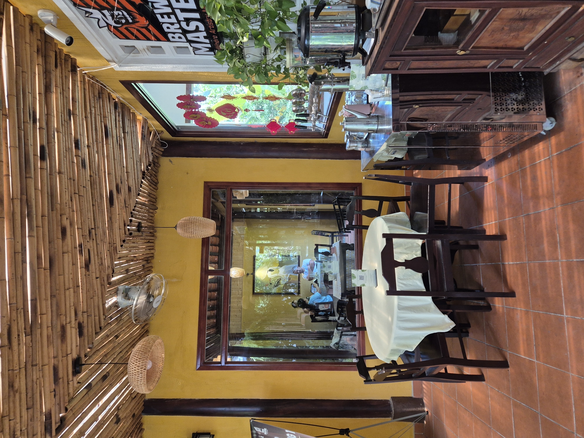 The image depicts the interior of a restaurant in Đà Nẵng, Vietnam, with a distinctly natural and rustic aesthetic. The ceiling is constructed from bamboo poles, and woven bamboo lamps hang from it. The walls are painted a vibrant yellow.
In the foreground, a round table is covered with a light-colored tablecloth and surrounded by dark wooden chairs. In the middle ground, a large window looks into another section of the restaurant, where two individuals are seated at a table. One person, wearing a blue shirt, appears to be looking at something on the table, while the other, in a pink shirt, is gesturing. A framed picture is visible on the wall behind them.
To the right of the window, a stainless steel bar area is equipped with beer taps and a large, silver kettle or urn. A dark wooden cabinet stands next to the bar. Green potted plants are arranged on a shelf above the bar area, and decorative red paper lanterns hang in the window. A sign with the words "BREW MASTER" and "Where Artistry..." is mounted on the wall. The floor is tiled with terracotta-colored square tiles. The overall atmosphere suggests a casual dining establishment with an emphasis on natural materials.