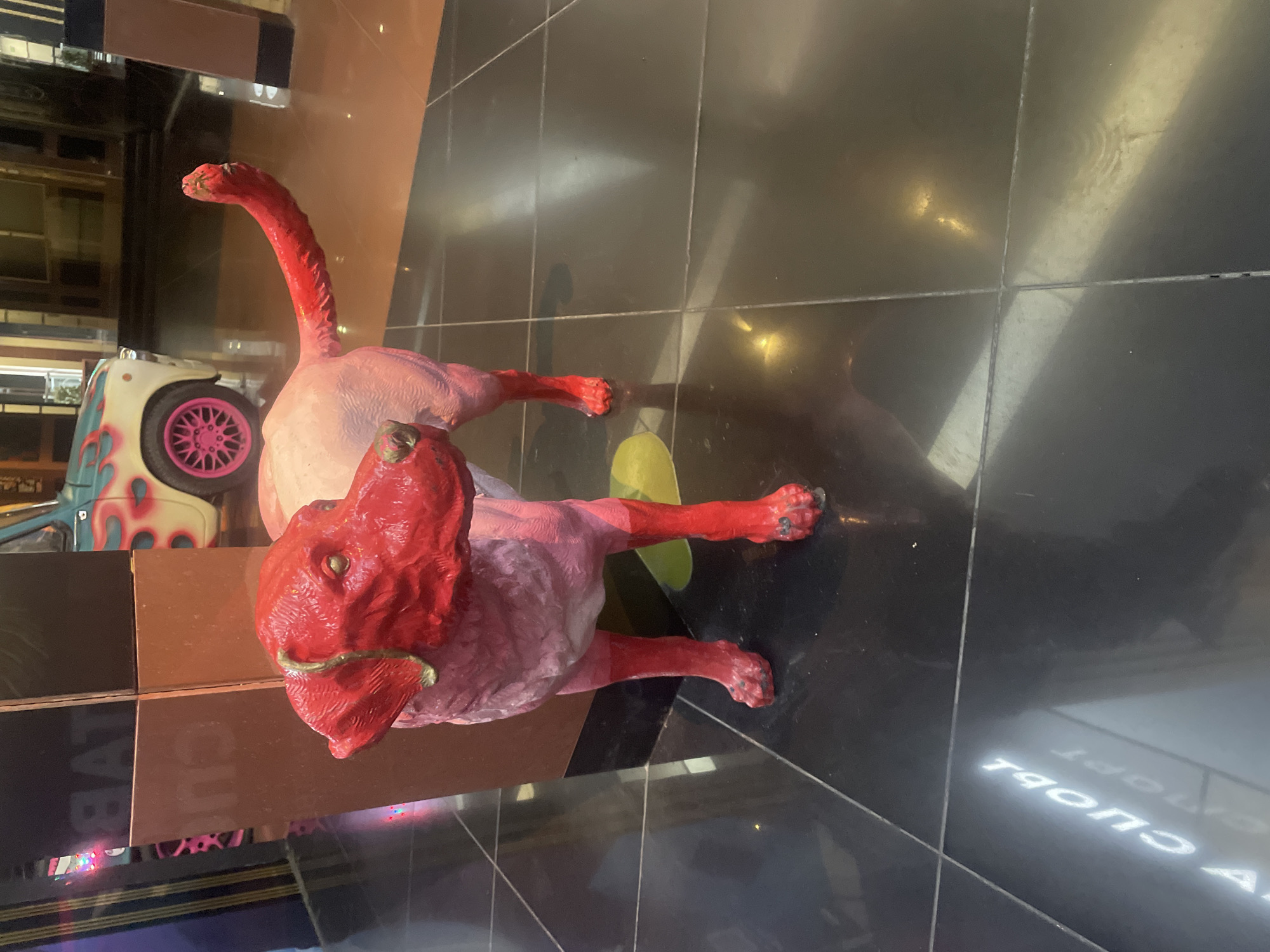 A stylized, vibrantly colored statue of a dog stands on a polished black tiled floor in what appears to be an indoor public space, possibly a shopping center or entertainment venue. The dog statue has a predominantly pink body with bright red accents on its head, ears, legs, and tail. The dog's head is tilted slightly, and its expression is neutral. Behind the dog, a small, retro-style car with flame decals is partially visible. Further in the background, there are architectural elements that suggest a commercial setting, including what appear to be display structures and possibly entrances or escalators. The lighting is artificial, casting reflections on the floor and creating a somewhat dramatic atmosphere. The overall impression is one of a modern art installation or a themed decorative element within a city.