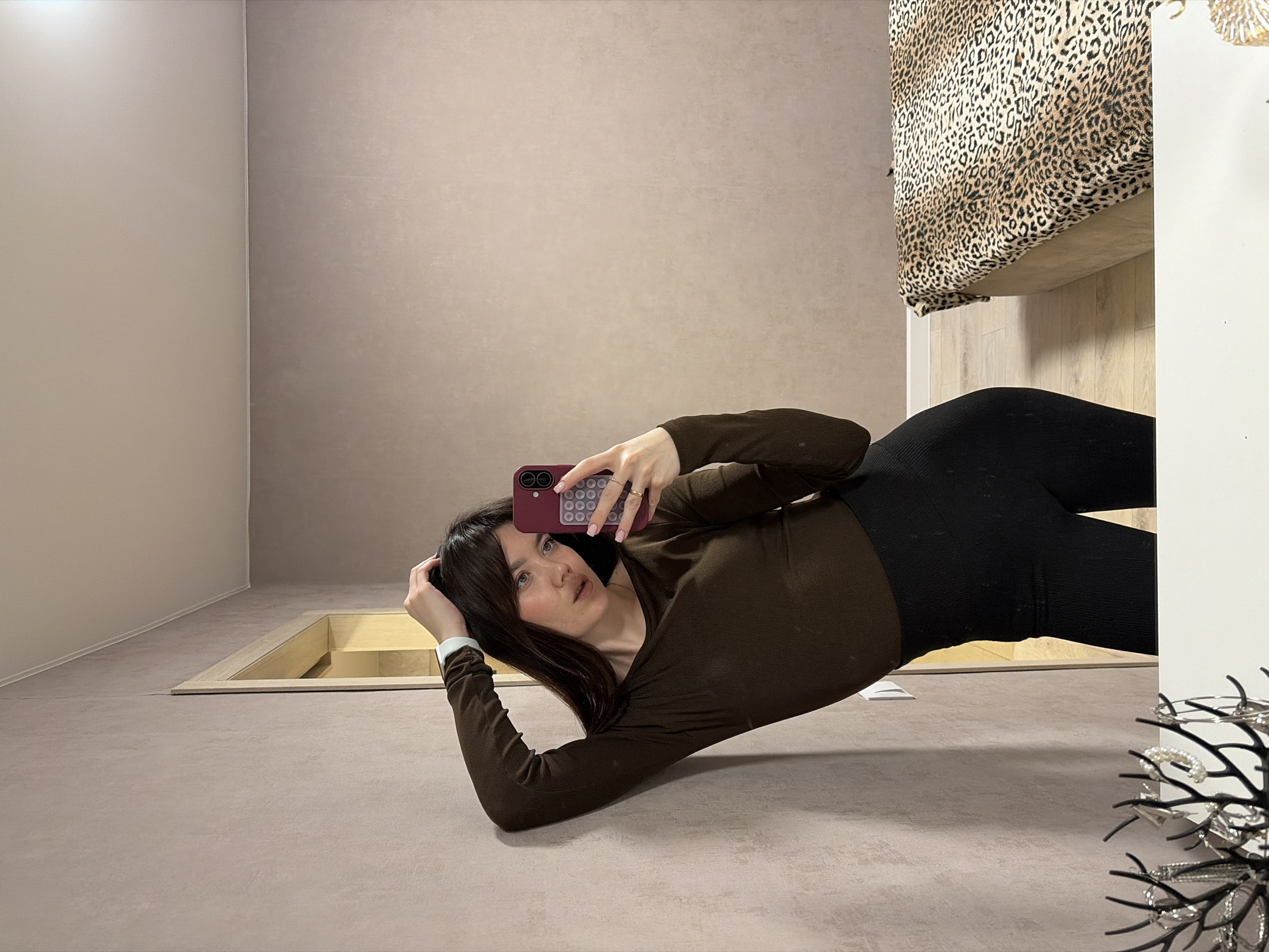 A woman is lying on a beige floor, taking a selfie with a maroon phone. She is wearing a brown long-sleeved shirt and black leggings. Behind her is a beige wall and what appears to be a sunken, rectangular light fixture in the floor. To her right, there is a bed with a leopard-print blanket. A white cabinet or wall and a jewelry stand are visible in the foreground. There is no indication of this photo's location in Votkinsk, Russia.