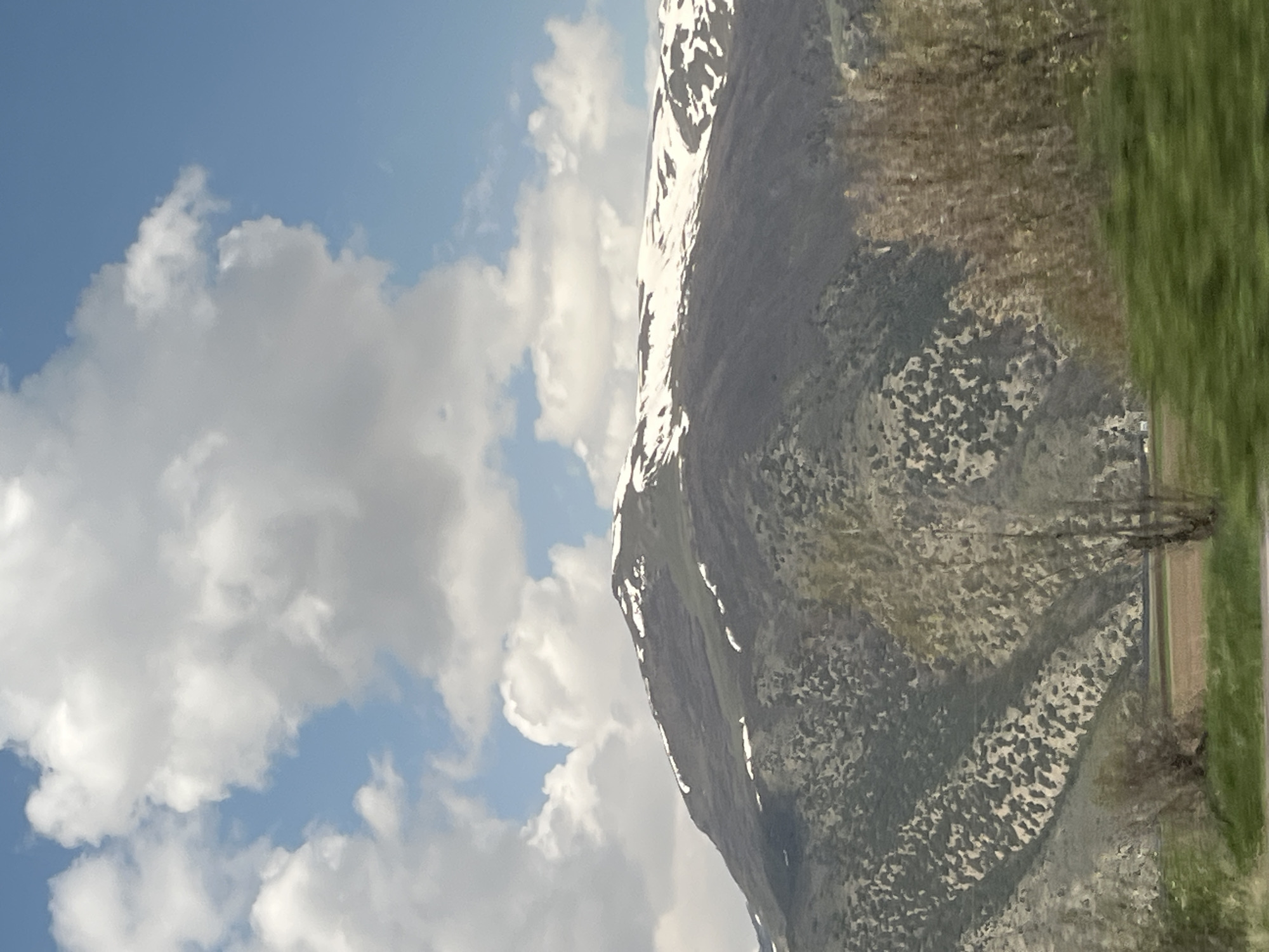The image shows a mountain partly covered in snow under a sky with scattered clouds. A road and some vegetation are visible in the foreground. The location is not explicitly mentioned in the image.
