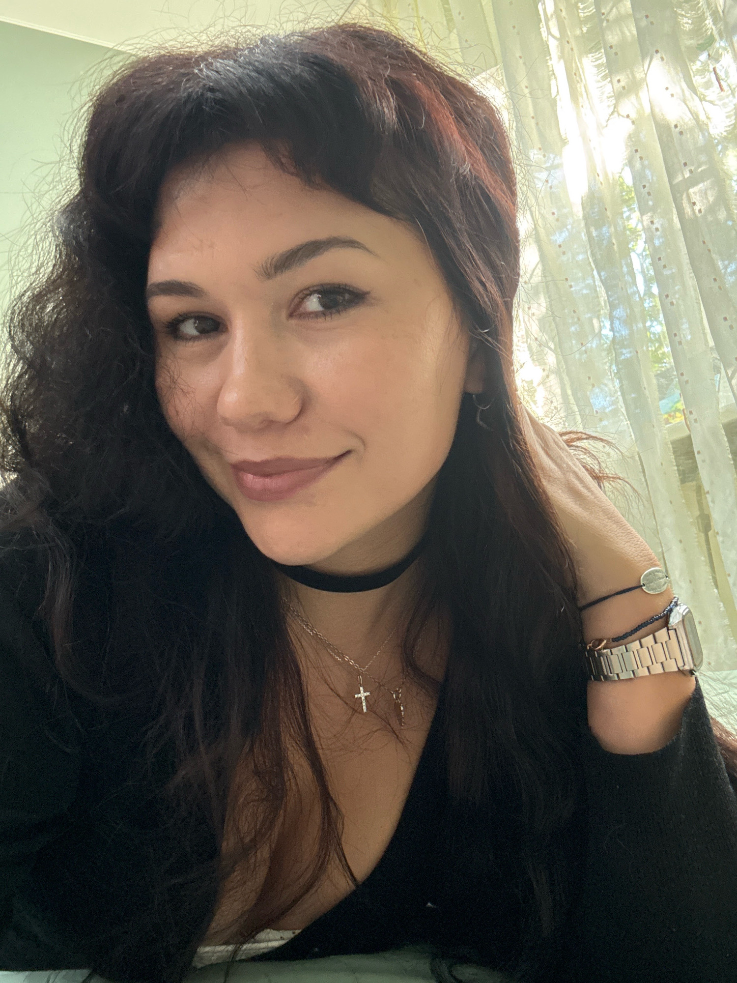 A woman with dark, wavy hair and bangs is shown from the chest up, looking slightly to her left and smiling. She is wearing a black top, a black choker necklace, and a silver necklace with a cross pendant. On her right wrist, she has a silver watch and a bracelet. She is standing in front of a sheer white curtain with a window behind it, through which sunlight is filtering, creating shadows. The background wall is painted a light green. There is no indication of the location in the city of Cheboksary, Russia.