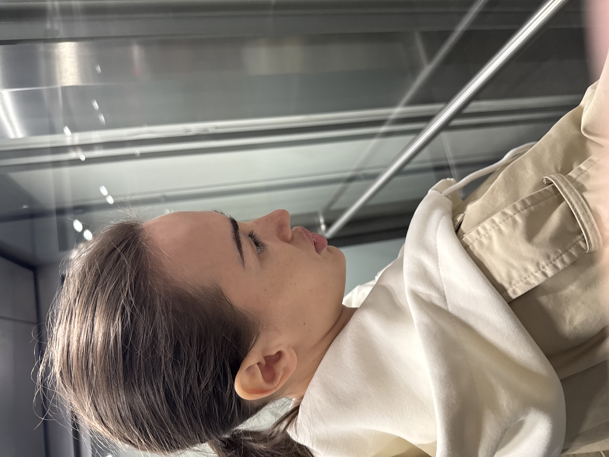 A woman with brown hair pulled into a low ponytail is lying down, facing towards the right of the frame. She is wearing a white hooded sweatshirt and tan pants with a visible pocket. She appears to be inside a modern elevator or similar structure with metal and glass surfaces. This cannot be definitively determined to be in Warsaw, Poland without additional evidence.