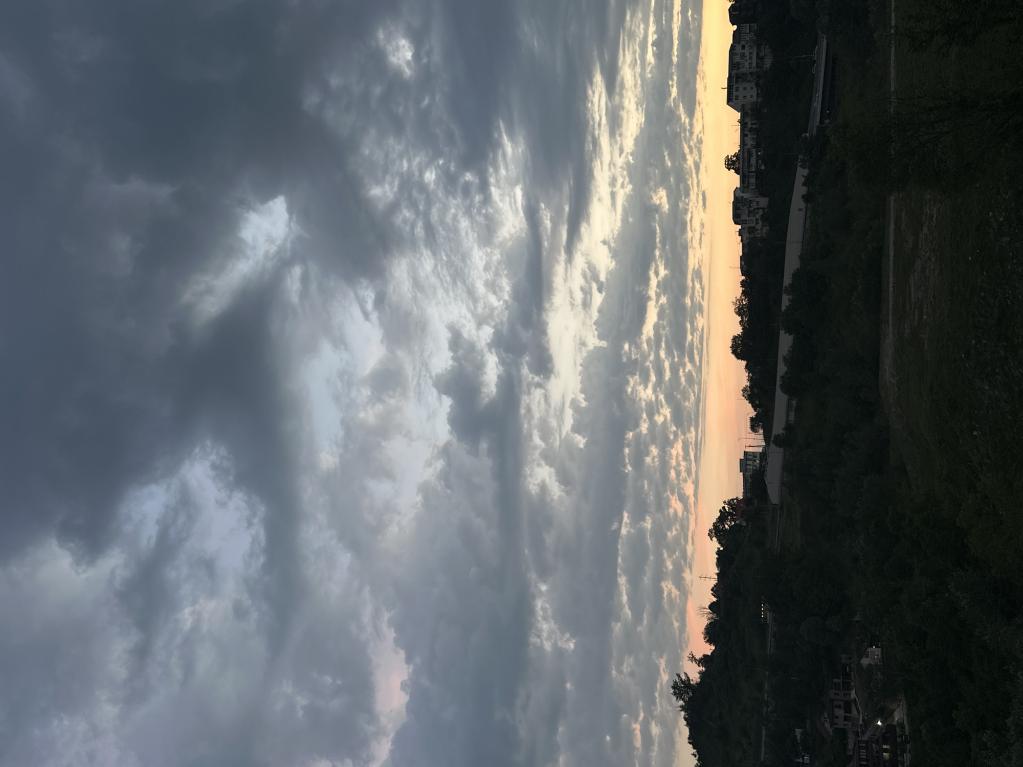 The image shows a view of a residential area on a hillside in Sochi, Russia, during a cloudy sunset. The sky is filled with dark, heavy clouds, with patches of lighter sky and some hints of orange and pink from the setting sun peeking through. Below the sky, a dense green hillside is covered in trees and vegetation. On the hillside, several buildings are visible, including multi-story apartment buildings and houses. A road or highway can be seen winding through the landscape. The overall impression is a slightly overcast and dramatic end to the day in a coastal city.