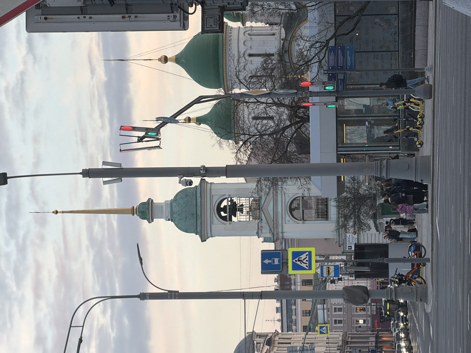 The image shows a street scene in Moscow, Russia. In the foreground, there are pedestrian crossings and traffic lights. Several people are waiting on the sidewalk. A few parked bicycles and scooters are also visible. In the background, several Russian Orthodox churches with green domes and golden spires can be seen. The sky is cloudy, and the light suggests it may be dusk or dawn. The urban environment is characterized by a mix of older religious architecture and modern city infrastructure.