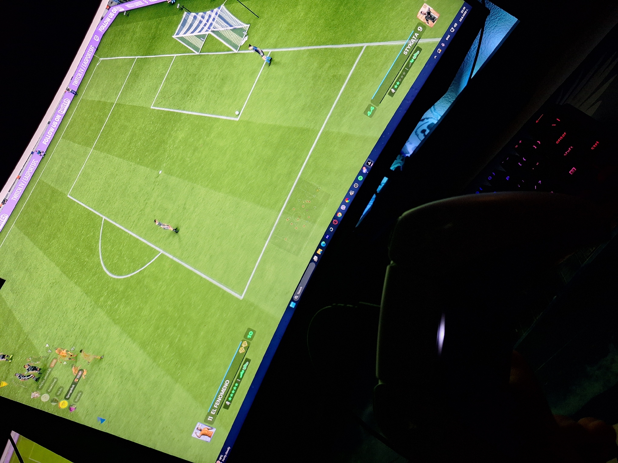 The image depicts a person playing a soccer video game, likely FIFA, on a large monitor. The game displays a soccer field with players and team information. In the foreground, a controller and a keyboard are visible, illuminated by the screen and the keyboard's RGB lighting. The monitor shows several player names and stats, such as "EL FENOMENO" and "STYKAТА 9". The background of the game screen has a banner with "TWITCH.TV/EASPORTSFC FOLLOW US ON TWITCH". The scene is dark, suggesting it is nighttime or the room is dimly lit, with the primary light source being the monitor. The image is taken from a high-angle perspective, looking down at the screen and the player's setup. There is no identifiable location in the city of Varna, Bulgaria, as the image only shows a digital environment and a player's gaming setup.