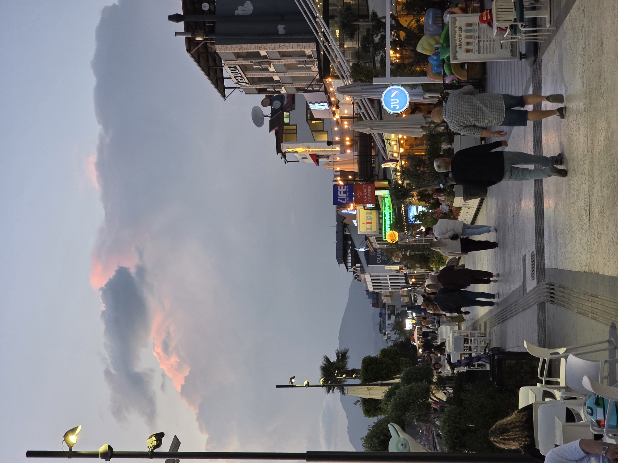 This image captures a vibrant street scene in Fethiye, Turkey, at dusk or early evening. The sky, on the left side of the frame, transitions from a muted grey-blue to a darker hue, punctuated by large, dramatic clouds tinged with soft pink and orange from the setting sun.
The right side of the image features a lively pedestrian street lined with multi-story buildings that house various businesses and possibly residences. Numerous people are visible, some walking along the paved street, while others are seated at outdoor tables and chairs, suggesting cafes or restaurants. Streetlights are illuminated, casting pools of warm light, and many shop signs glow with artificial light, contributing to the evening ambiance. Large white outdoor umbrellas are set up over some seating areas.
In the background, dark, imposing mountains rise against the twilight sky, hinting at the geographical setting. A street pole with multiple light fixtures is prominently positioned in the foreground on the lower left.
Visible text on signs includes "JU" within a circular blue sign, "LIFE," and "TERRACE COFFEE." Parts of other signs like "LIDL" and "OLIM" (likely OLYMPOS) are also discernable. A menu board displays "FAVORILER," and a building in the mid-ground bears the name "AZAGAROV" on its facade. The overall atmosphere is bustling and active, typical of a town center or promenade as night falls.