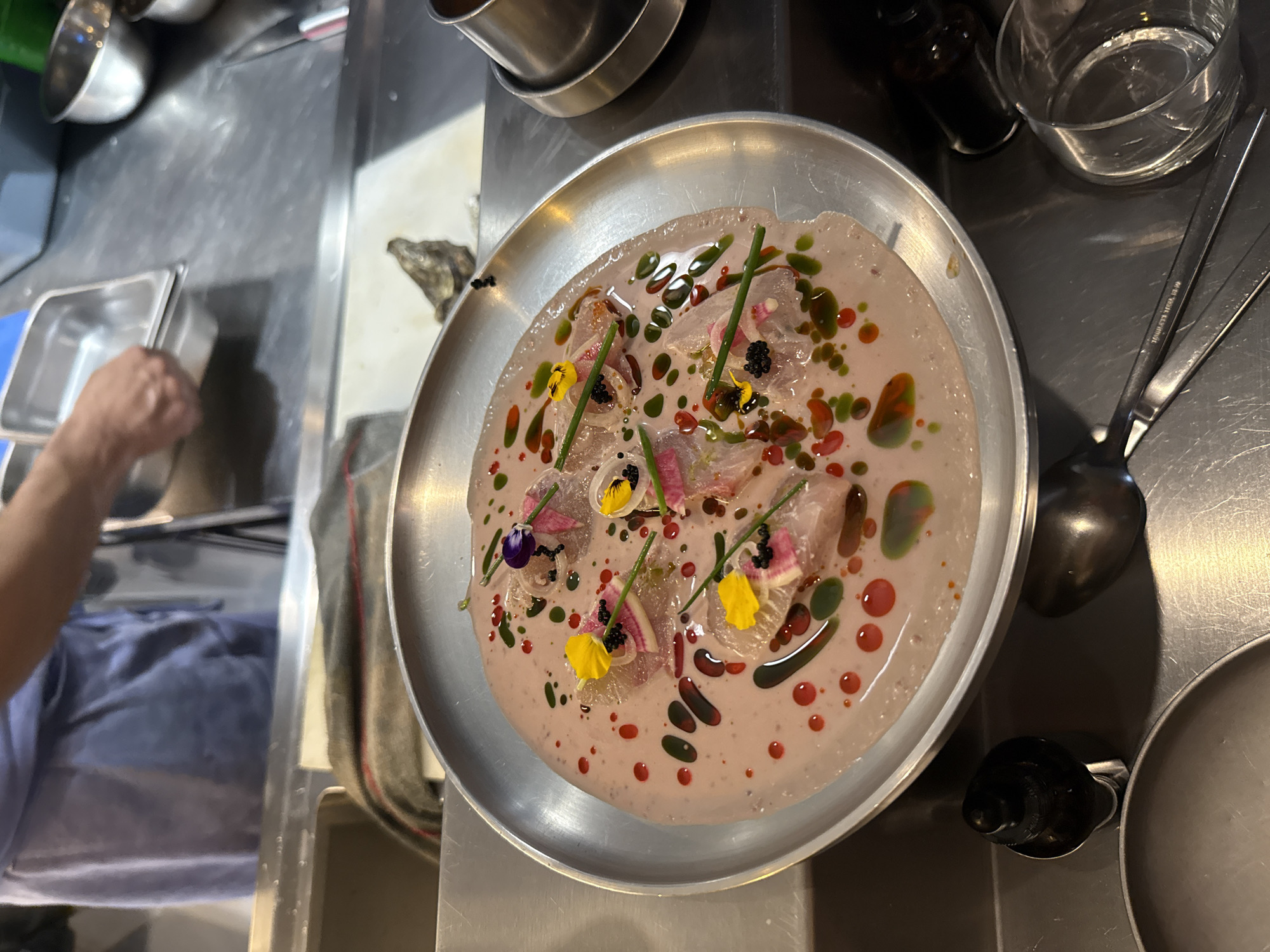 The image captures a gourmet dish, likely a carpaccio or ceviche variant, served on a silver platter. The dish consists of thinly sliced fish, radishes, edible flowers, chives, and what appears to be black caviar, all immersed in a pale pink sauce speckled with dots of red and green oils. The plate is placed on a metallic kitchen counter, with various culinary tools and containers visible in the background, suggesting a professional kitchen environment. A person's hand can be seen on the left of the picture, likely preparing the dish. The image does not provide information to confirm this location is Mexico City, Mexico.