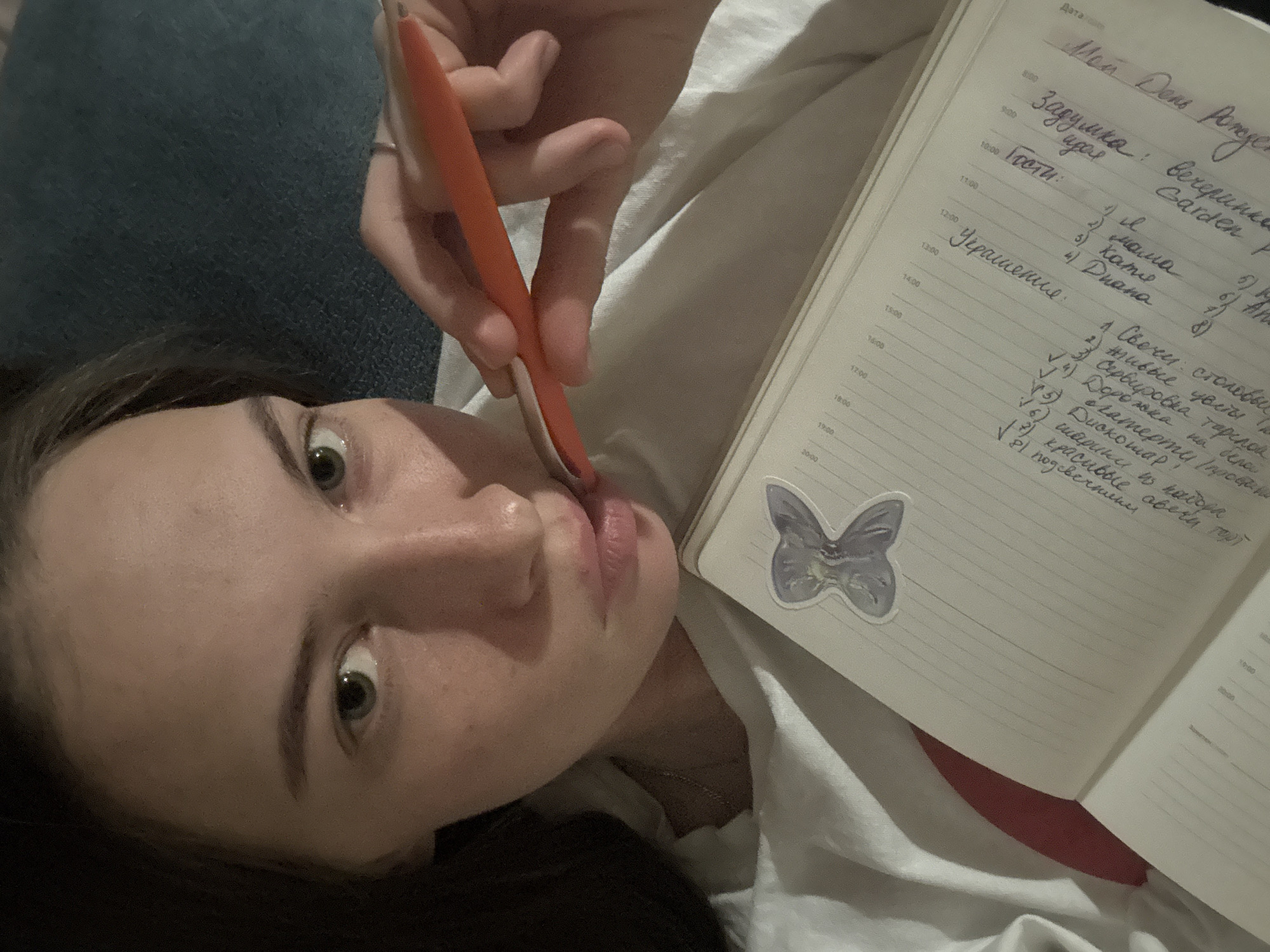 A young woman is lying down and looking at the camera, holding an orange pen to her lips. She is wearing a white t-shirt. In front of her is an open notebook with handwritten text. There is a sticker of a purple butterfly on the notebook. The environment appears to be indoors, possibly a bedroom or living room, with a blue textured surface in the background. No information is present to indicate the city of Perm, Russia.
