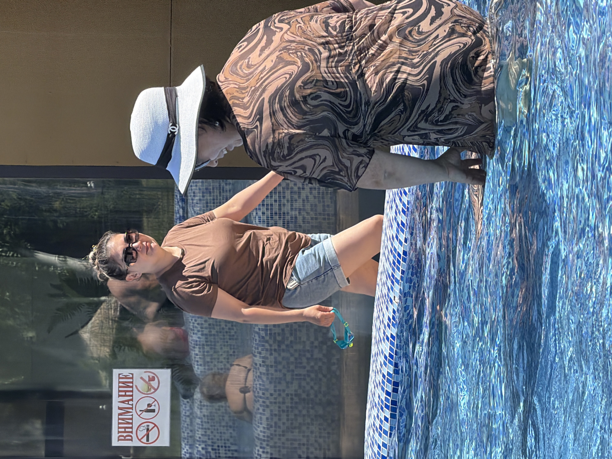 Two women are at the edge of a swimming pool on a sunny day. The woman on the left is wearing a brown t-shirt and denim shorts, and is holding a pair of goggles. She is looking towards the left side of the frame. The woman on the right is wearing a patterned dress and a white hat, and is facing away from the camera. She is smiling and her hand is in the water. In the background, through a glass panel, other people can be seen in what appears to be a hot tub or spa area. There is a sign with Russian text and no-smoking, no-drinking, and no-eating symbols on the glass panel. The pool is tiled with blue and white mosaic.