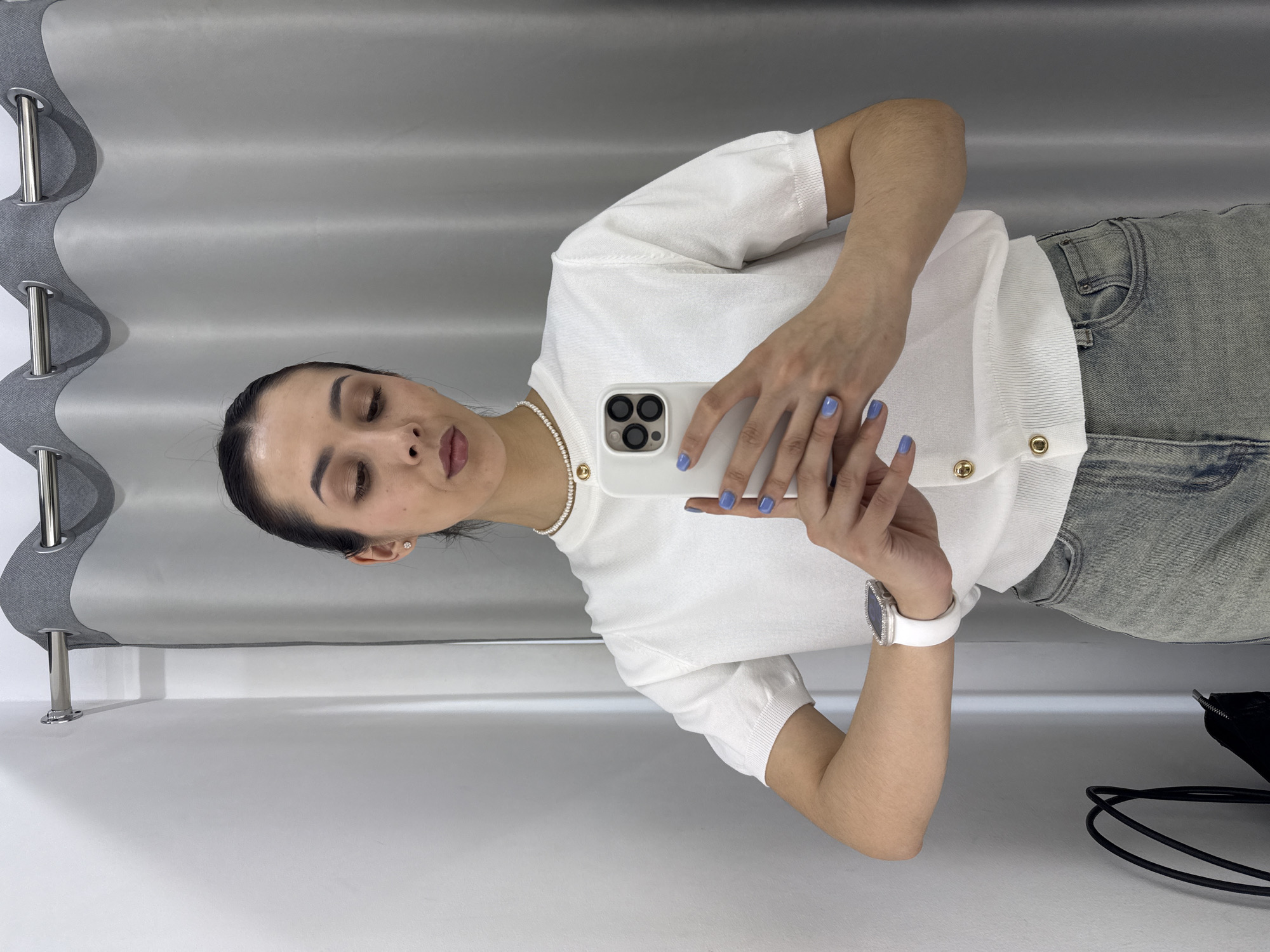 A woman is taking a selfie indoors in a clothing store setting. She is holding a white smartphone with three lenses in the upper left corner of the phone. She is wearing a white short-sleeved top with gold buttons, light blue jeans, a white watch, and a pearl necklace. Her nails are painted a light blue color. The background includes a gray curtain and a white wall. The location is likely a fitting room in a clothing store in Votkinsk, Russia.