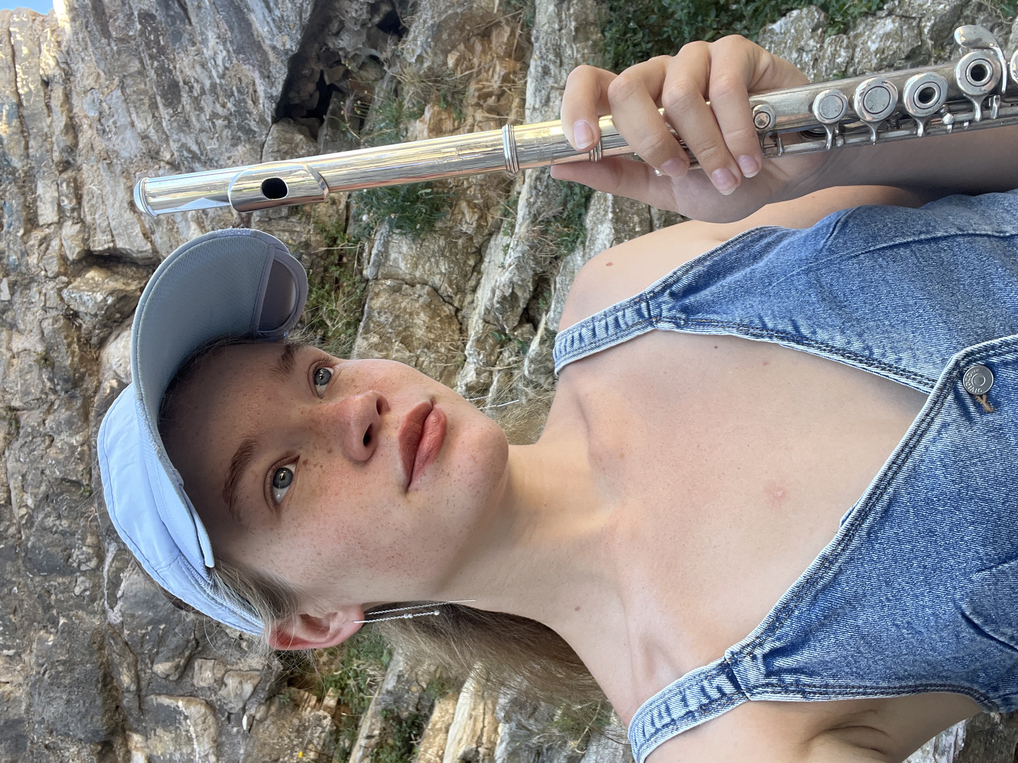 A young woman with freckles and blonde hair pulled back stands outdoors, holding a silver flute. She is wearing a light blue baseball cap and a denim sundress. The background is a rocky cliff face with some greenery. The image appears to be a selfie, taken outdoors during the day. The location is the city of Budva, Montenegro, near the coast.