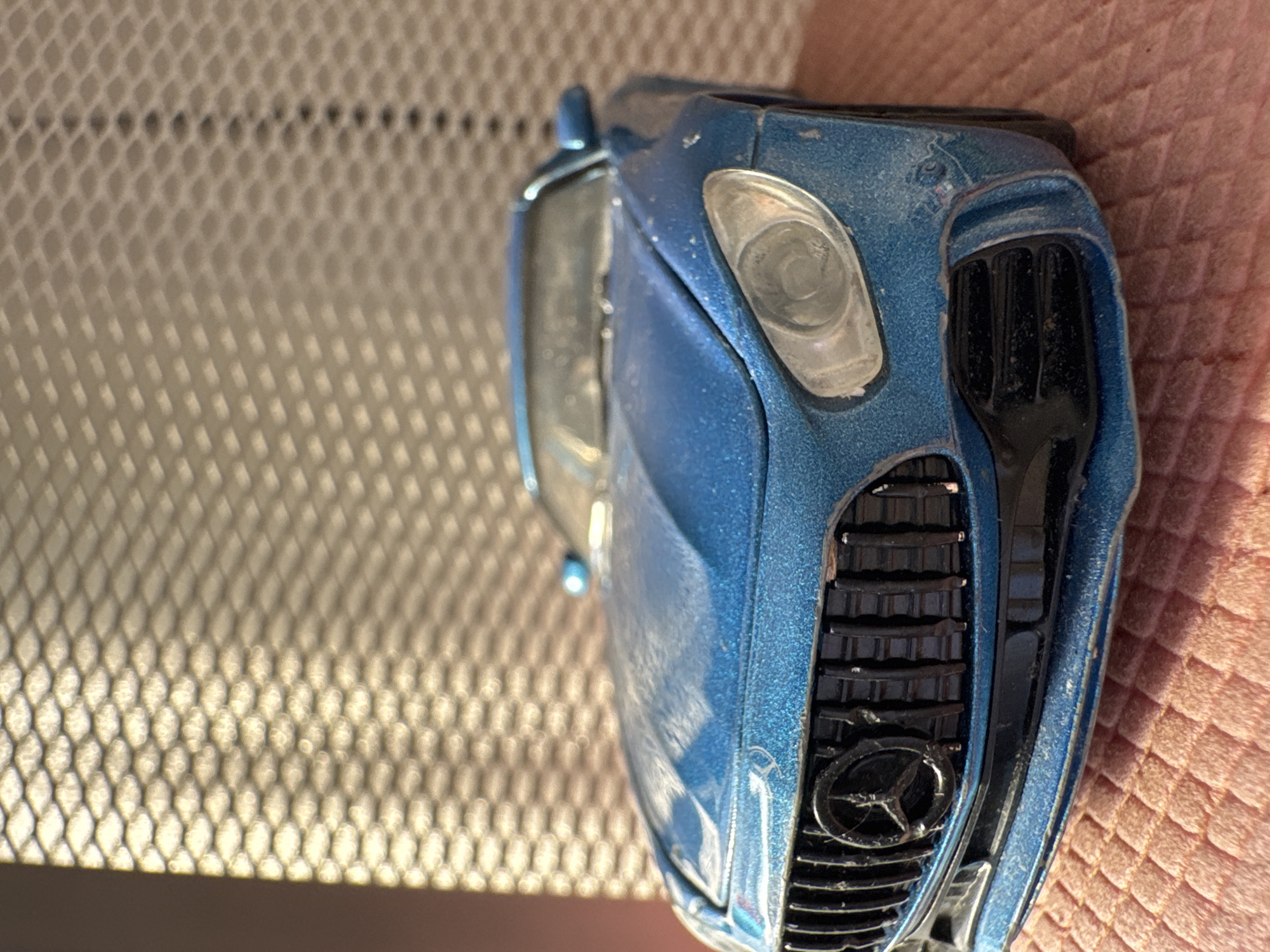A close-up shot depicts the front of a blue toy car, specifically the grill, headlight, and part of the hood. The car is situated on what appears to be a surface with a patterned texture, possibly a woven material or mesh. The background is a combination of light and shadow, adding depth to the image. No people, actions, or interactions are visible. There is no information to indicate that this scene is taking place in Novo-Nikol'skoe, Russia.