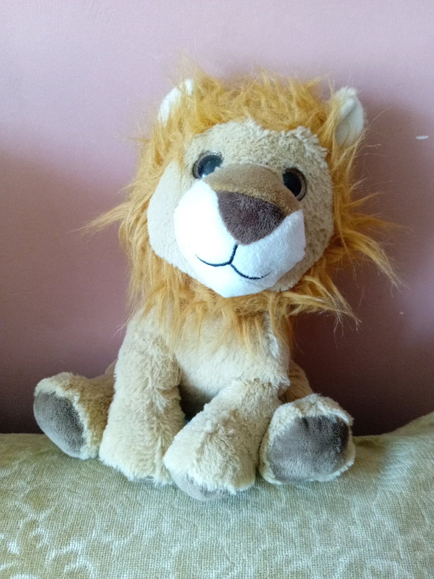 A stuffed toy lion with a shaggy, orange mane sits on a textured, light-colored surface against a pink wall. The lion is light brown with a white muzzle, a brown nose, and black button eyes. Its mouth is depicted by a black stitched line. The toy is positioned centrally in the frame, with its paws resting on the surface. The background is a plain pink wall, and the foreground is occupied by the textured surface on which the toy is placed. There are no people present in the image, and no specific location in Tiachiv, Ukraine, is identifiable from the scene.