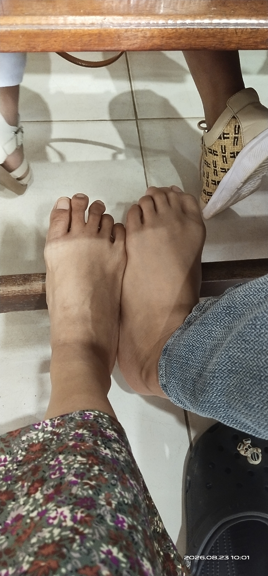 The image shows a close-up view of two bare feet positioned on a tiled floor. The feet are positioned side-by-side, with the toes of both feet visible. One foot is closer to the camera and is slightly angled. The other foot is mostly obscured by the first foot and the leg of a person wearing blue jeans. The environment appears to be indoors, possibly under a wooden table, as indicated by the texture and color of the surface in the upper part of the image. Shadows are cast on the tiled floor, suggesting a light source from above or the side. A part of a shoe with a patterned design is visible on the right side of the image, and another shoe, black and open-toed, is partially visible at the bottom right corner. A floral-patterned garment is visible on the left side, covering a leg. The timestamp "2026.08.23 10:01" is present at the bottom right of the image. There is no information about the city of Jaar, Indonesia in the image.