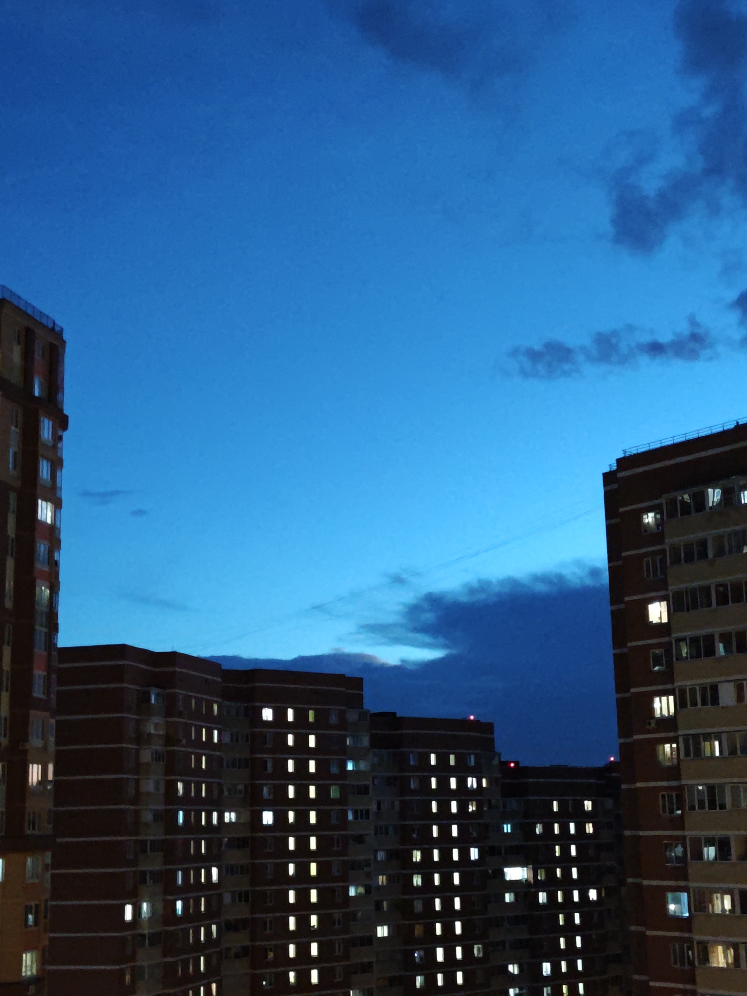 This media file depicts a twilight view of apartment buildings in Shcherbinka, Russia. The sky is a deep blue, transitioning from the day's end, with a few scattered dark clouds. The primary subjects are multi-story residential buildings, characterized by their brick and light-colored facades. Many windows are illuminated, indicating that residents are inside their homes, suggesting a typical evening in a residential area.
The activity taking place is simply the existence of urban life at dusk, with the lights in the apartments signifying habitation and daily routines. There are no visible people or specific events captured. Notable details include the time of day being twilight or early evening, the weather appearing partly cloudy, and the location being identifiable as Shcherbinka, Russia. The mood is serene and perhaps a little melancholic, typical of a fading day in a large residential complex. The illuminated windows create a pattern across the facades of the buildings, adding a sense of order and human presence to the scene. No text is visible in the image.