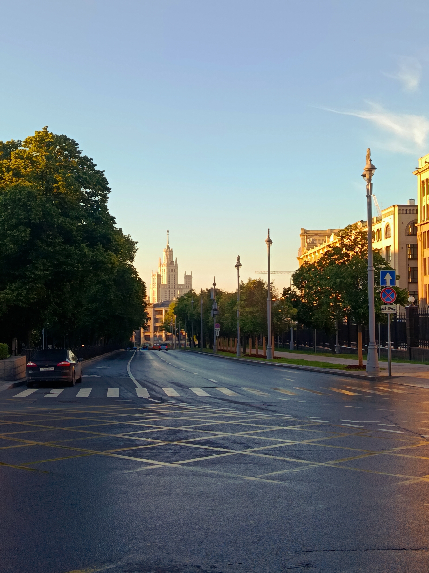 This is a daytime, eye-level, outdoor, medium shot of a city street with a clear sky. A wet, paved road occupies the foreground and extends into the background, with yellow crosswalk markings and diagonal lines visible on the asphalt. On the left side of the road, a dark-colored sedan is parked, partially obscuring the pedestrian crosswalk. Tall, lush green trees line the left side of the street, their canopies creating shade. On the right side, a row of streetlights and a fence separate the road from buildings. The buildings appear to be constructed of light-colored brick or stone. In the distance, a tall, ornate skyscraper with a spire dominates the skyline. The lighting suggests either early morning or late afternoon, with warm sunlight casting a glow on the scene. The overall impression is one of a peaceful urban setting during a clear day.