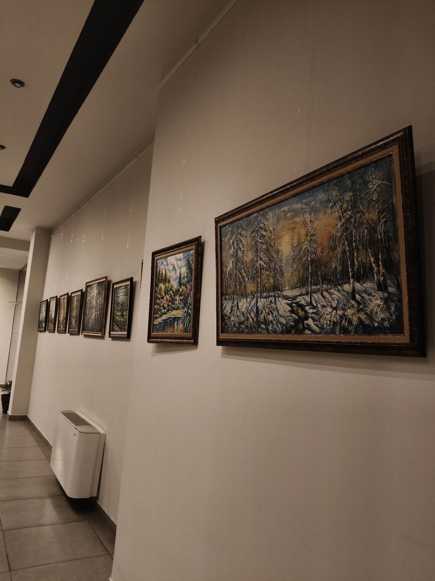 A hallway in Plovdiv, Bulgaria, features a series of framed landscape paintings hanging on a white wall. The paintings appear to depict scenes of forests and nature, some with snow. A white radiator is visible on the floor near the base of the wall. The hallway extends into the distance. The ceiling has a decorative black stripe. The paintings are hung using clear hanging wires.
