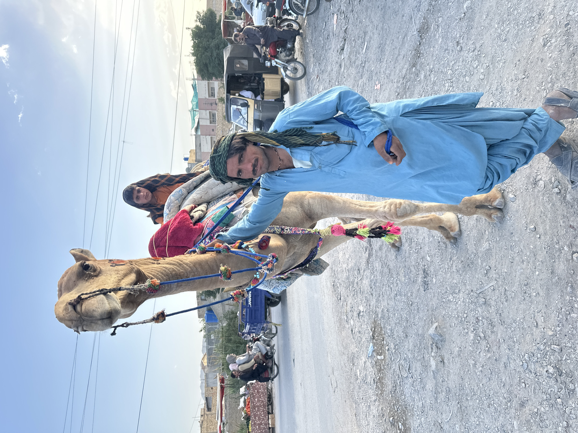 A camel stands on a road in Quetta City Tehsil, Pakistan. A man in a blue outfit and headscarf stands next to it, smiling at the camera while holding a blue string connected to the camel. On the camel's back, partially hidden behind a blanket, is another person with a brown shawl. The camel is decorated with colorful ropes. In the background, there are buildings, vehicles, and other people. The sky is partly cloudy.