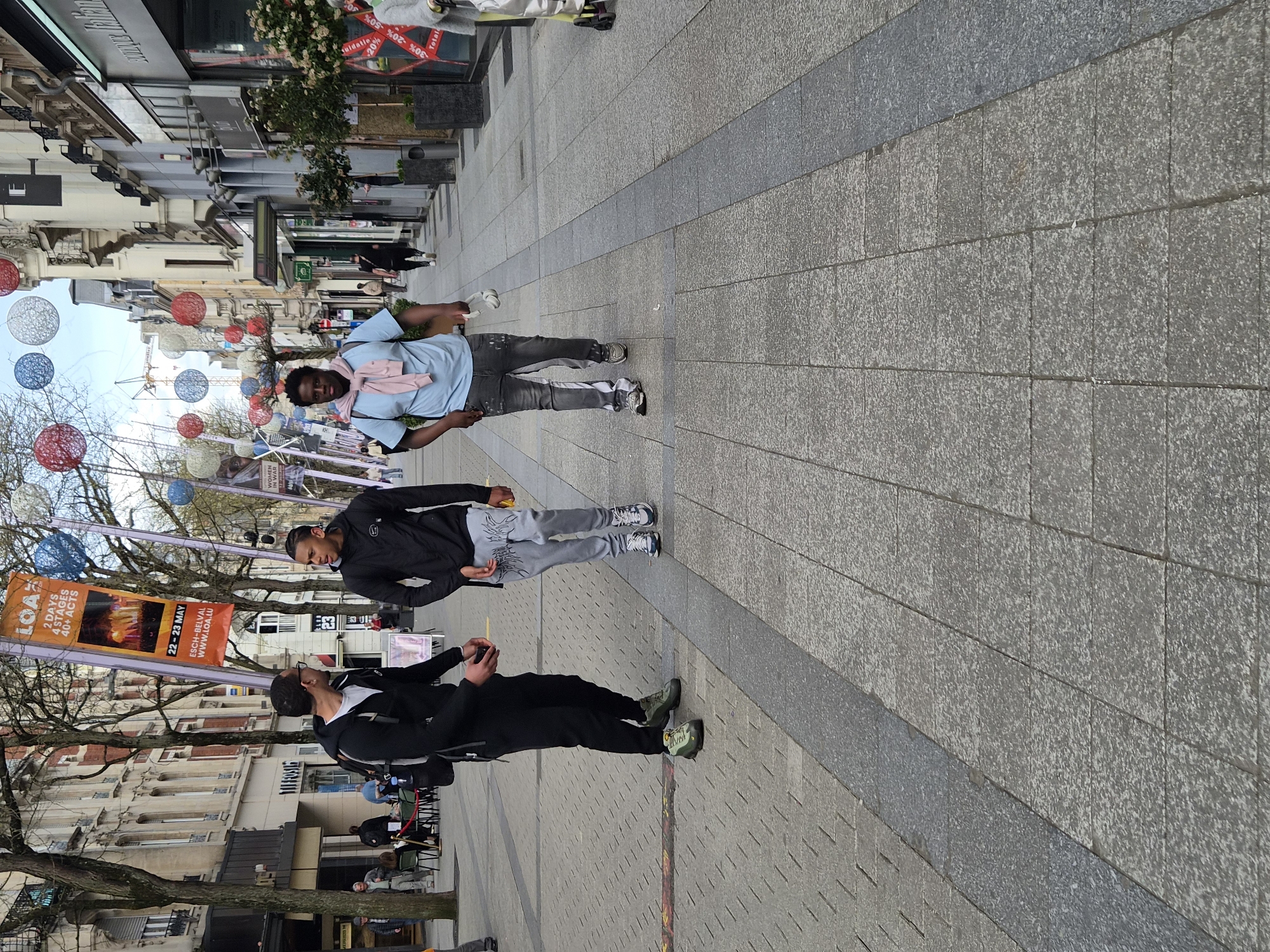This daytime image captures a pedestrian street in Esch-sur-Alzette, Luxembourg, adorned with decorative spherical lanterns in red, white, and blue. The street is paved with light and dark grey blocks, flanked by multi-story buildings that appear to house shops. Leafless tree branches line the street.
In the foreground, three men are visible. Closest to the camera on the right, a man with dark hair and a beard wears black clothing and green and black athletic shoes, holding a cup-like object. To his left, another man in a black jacket, patterned light grey sweatpants, and athletic shoes looks towards the right. Further back, a third man wears a light blue shirt, a pink scarf, dark jeans with paint splatters, and athletic shoes, holding a white item. Other indistinct figures are visible further down the street and in the background.
Notable text includes a large orange banner promoting an event: "LOAZ," "2 DAYS 4 STAGES 40+ ACTS," scheduled for "22 - 23 MAY" in "ESCH-BELVAL," with the website "WWW.LOA.LU." Storefront windows display sale signs with percentages like "% -30% -20% -50%" and the word "Totald." Another sign partially visible reads "WOMEN." The scene depicts a casual, unhurried urban setting, likely a commercial or public square.