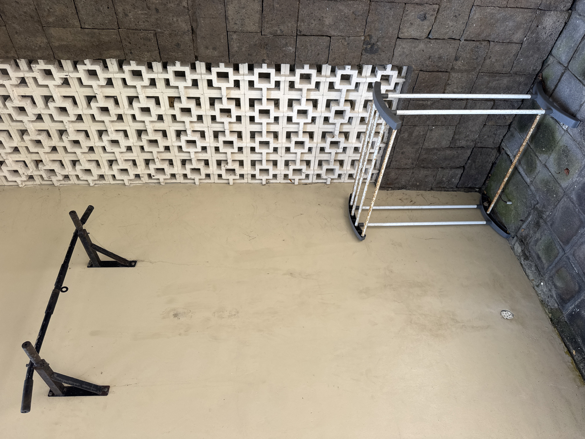 The image shows an outdoor space in Jimbaran, Indonesia. The ground is a light beige cement. A black, wall-mounted pull-up bar is attached to the left wall. A white metal clothes drying rack stands in the right corner. The walls are constructed of concrete blocks, with a decorative white block wall separating the beige wall from a darker gray stone wall above. There are no people visible.
