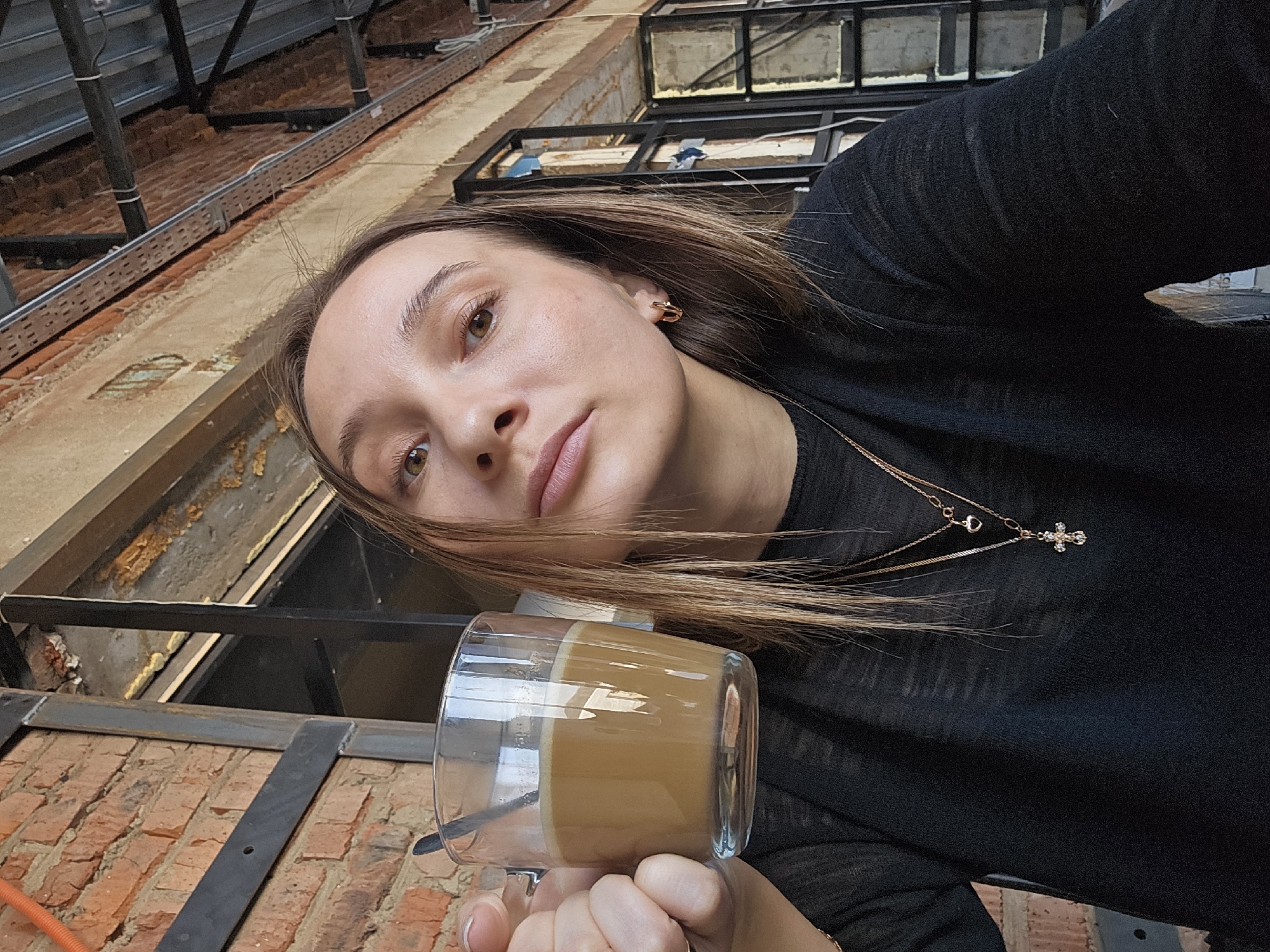 This image depicts a young adult female in an industrial-style interior located in Chaikovsky, Russia. The female, possessing light skin, brown eyes, and long light brown hair, is wearing a dark, fine-knit long-sleeved top. She accessorizes with two gold-toned necklaces, one featuring a heart charm and the other a longer chain with a clear-stoned cross pendant, along with small gold-toned hoop earrings. A thin gold-toned ring is visible on her left ring finger, and her fingernails are painted white. She holds a clear glass mug in her left hand, containing a creamy, light brown beverage, consistent with coffee or latte, and a silver-colored spoon. Her gaze is directed towards the camera. The background features exposed red brick walls, dark metal structural beams, exposed ceiling elements, and window frames. Remnants of insulation, specifically spray foam, and wiring are visible among the structural components.