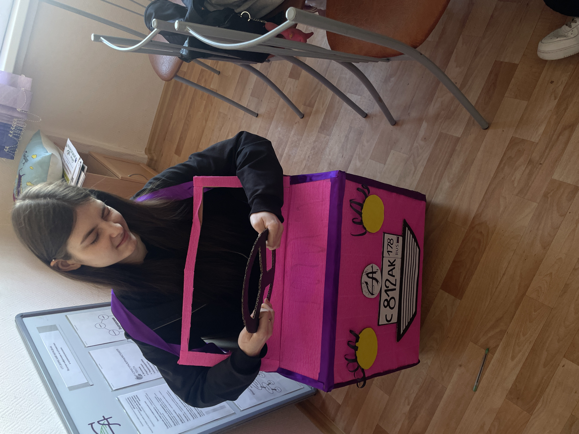 A woman is wearing a pink and purple cardboard box designed to look like a car, complete with a steering wheel, headlights, and a Russian license plate that reads "C 812 AK 178 RUS." The "178" number on the plate indicates that this is the city of Saint Petersburg in Russia. She is standing in what appears to be a classroom with wooden floors. There is a whiteboard with papers attached behind her, and chairs and a table are in the background.
