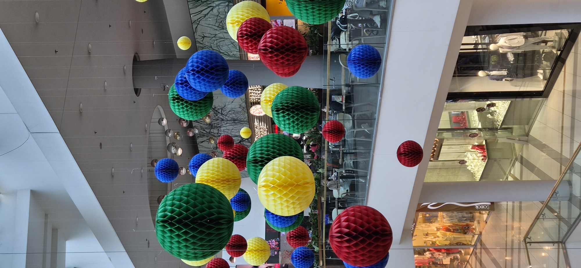 The image shows an interior view of a multi-level shopping mall in Varna, Bulgaria. Hanging from the ceiling are numerous colorful honeycomb paper spheres in red, yellow, green, and blue. These are suspended by thin, clear strings. The ceiling above is a gray, modern design with recessed lighting and architectural lines. Shopfronts with glass displays are visible on multiple levels, reflecting the surrounding environment. Mannequins are dressed in clothing inside the shops. The overall environment suggests a commercial space designed to attract customers with vibrant decorations.