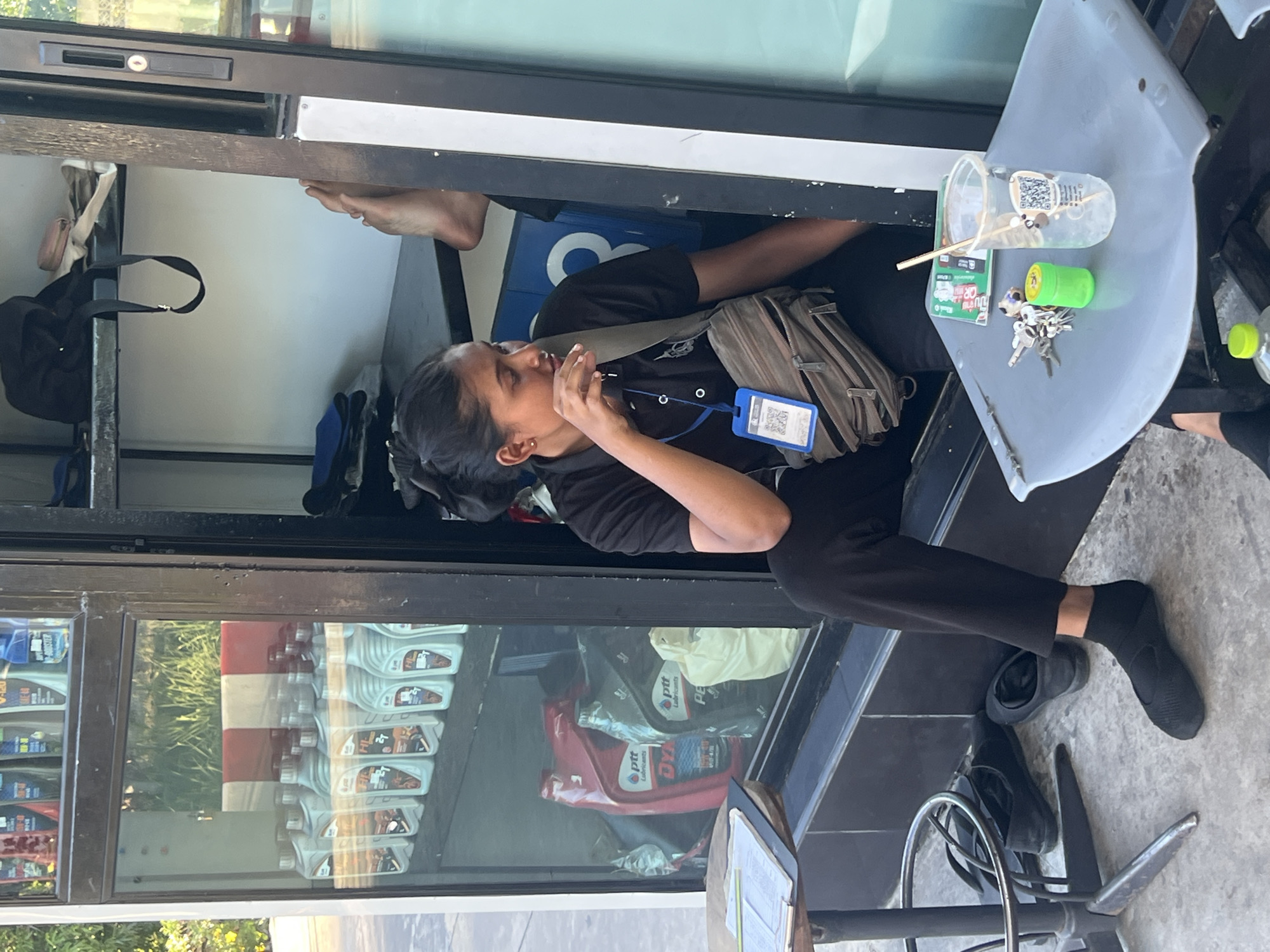 A person wearing a black shirt and black pants is leaning out of a window or service counter. They have a light brown crossbody bag and an identification card with a QR code clipped to their shirt. The person appears to be talking or gesturing with their hand near their mouth.
On the counter, there is a clear plastic cup with a straw and a QR code, a small green object, and a set of keys. Behind the person, shelves are visible with various items, including what looks like bottles of oil or lubricant.
The scene is outdoors, with natural light suggesting daytime. The reflection in the window shows greenery and a red and white striped structure, possibly part of a building or display. The overall setting appears to be a shop or service point, with products displayed on shelves. The ground is paved. The location is identified as Koh Phangan, Thailand.