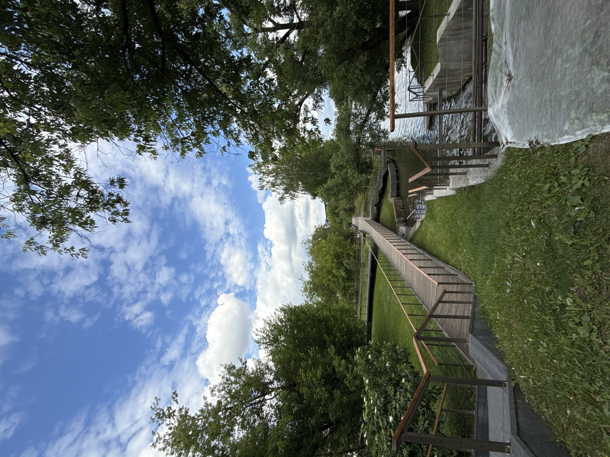 The image depicts a wooden walkway with metal railings leading towards a body of water, likely a river or lake, in Moscow, Russia. The walkway is surrounded by lush green grass and mature trees, with a bright blue sky dotted with fluffy white clouds overhead. The water features a ripple effect, suggesting a gentle current or perhaps a small waterfall. In the distance, hints of urban structures, possibly buildings, can be observed through the trees. The scene appears to be a tranquil park or recreational area, designed to allow visitors to traverse the landscape and access the water's edge. The presence of a protective white covering over a section of vegetation on the right side of the image suggests ongoing landscaping or horticultural activity. There are no people clearly visible in the scene.