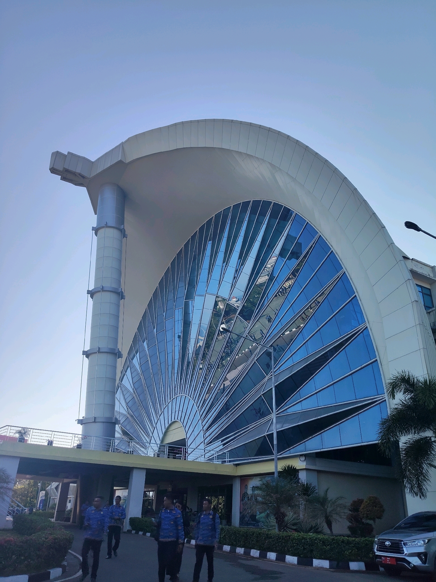 The image shows the exterior of a modern building with a large, fan-shaped glass facade. The building has a white, curved roof supported by a large, cylindrical pillar. The glass facade reflects the blue sky and surrounding environment, including what appears to be a street with cars and people. In the foreground, several men in matching blue batik shirts and black pants are walking on a paved area. Lush green bushes and palm trees are visible around the building. A silver SUV is parked on the right side of the frame. The scene appears to be set in a well-lit outdoor environment, likely during the day. The architecture suggests a public building, potentially a government or cultural center. The location is identified as the International Convention Center (ICC) in Kupang, Indonesia.