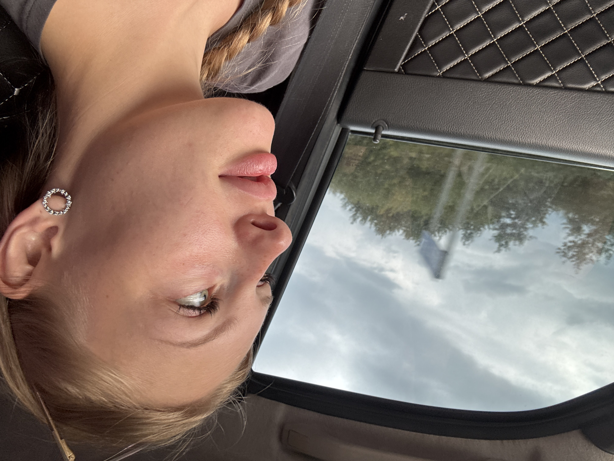 A young Caucasian woman with blonde hair in a braid is sitting in the passenger seat of a car, looking out the window. The interior of the car is visible, with a quilted black leather door panel and a gray seatbelt. Outside the window, there are trees and a cloudy sky. A blurry object resembling a flag on a pole is visible in the distance. The image was taken in Shumikha, Russia.