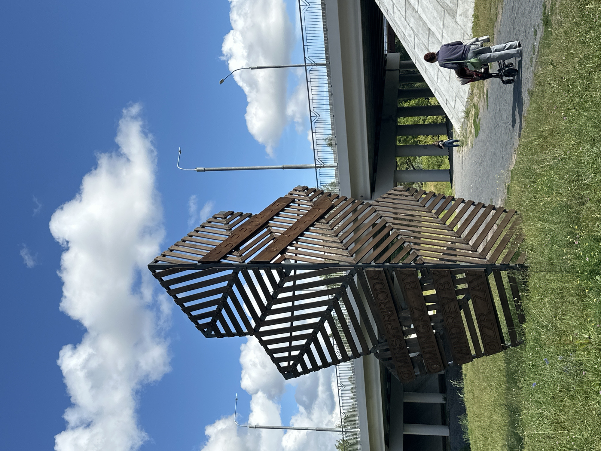 The image depicts an outdoor scene on a sunny day with a bright blue sky and scattered white clouds. In the foreground, a tall, abstract sculpture made of dark metal and wooden slats stands on a grassy area. The sculpture has a complex, geometric form. Attached to the wooden slats are several words that appear to be social media handles or names.
To the right of the sculpture, a paved walkway runs through the grass. A woman is pushing a stroller along this walkway. Further down the walkway, a child is walking away from the camera. In the background, a large concrete overpass or bridge spans across the scene, supported by concrete pillars. Streetlights are visible on the overpass. The area appears to be a park or public space, possibly near a road or infrastructure. The environment is green and appears well-maintained.