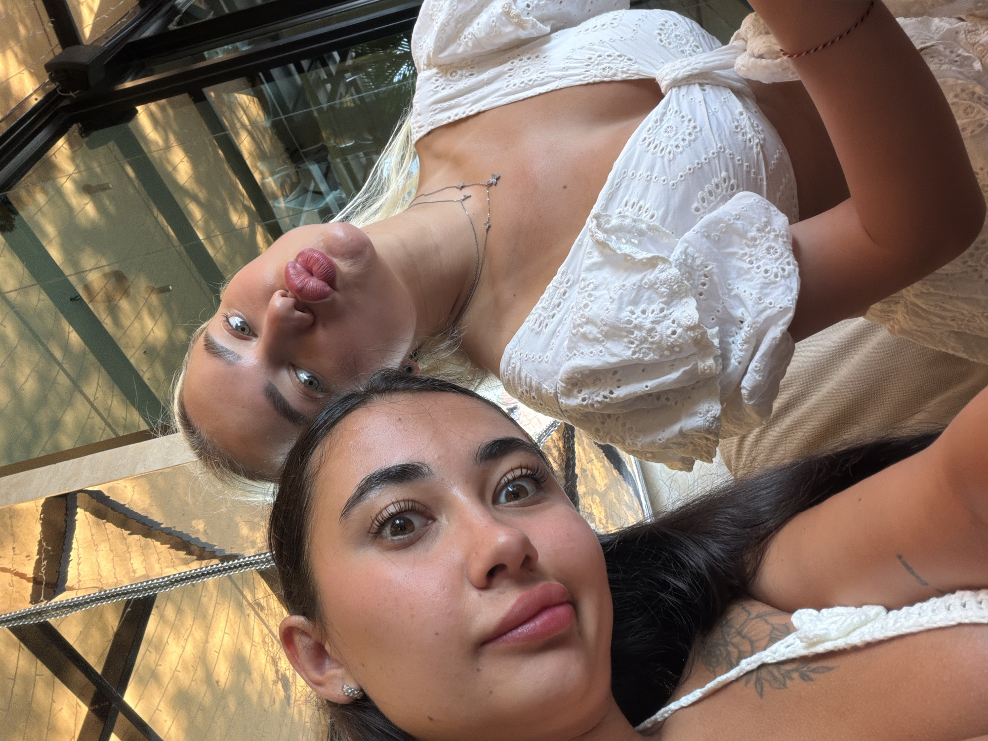 Two young women are posing for a selfie. The woman in the foreground has dark hair and brown eyes. She is wearing a white top with a crocheted strap. The woman behind her has blonde hair and grey eyes. She is wearing a white, eyelet-style blouse with puffed sleeves and a delicate necklace. They are both making a kissing face. The background features a modern glass and metal structure with reflected light patterns. The location appears to be inside a building with architectural details consistent with high-end resorts in Choeng Thale, Thailand.