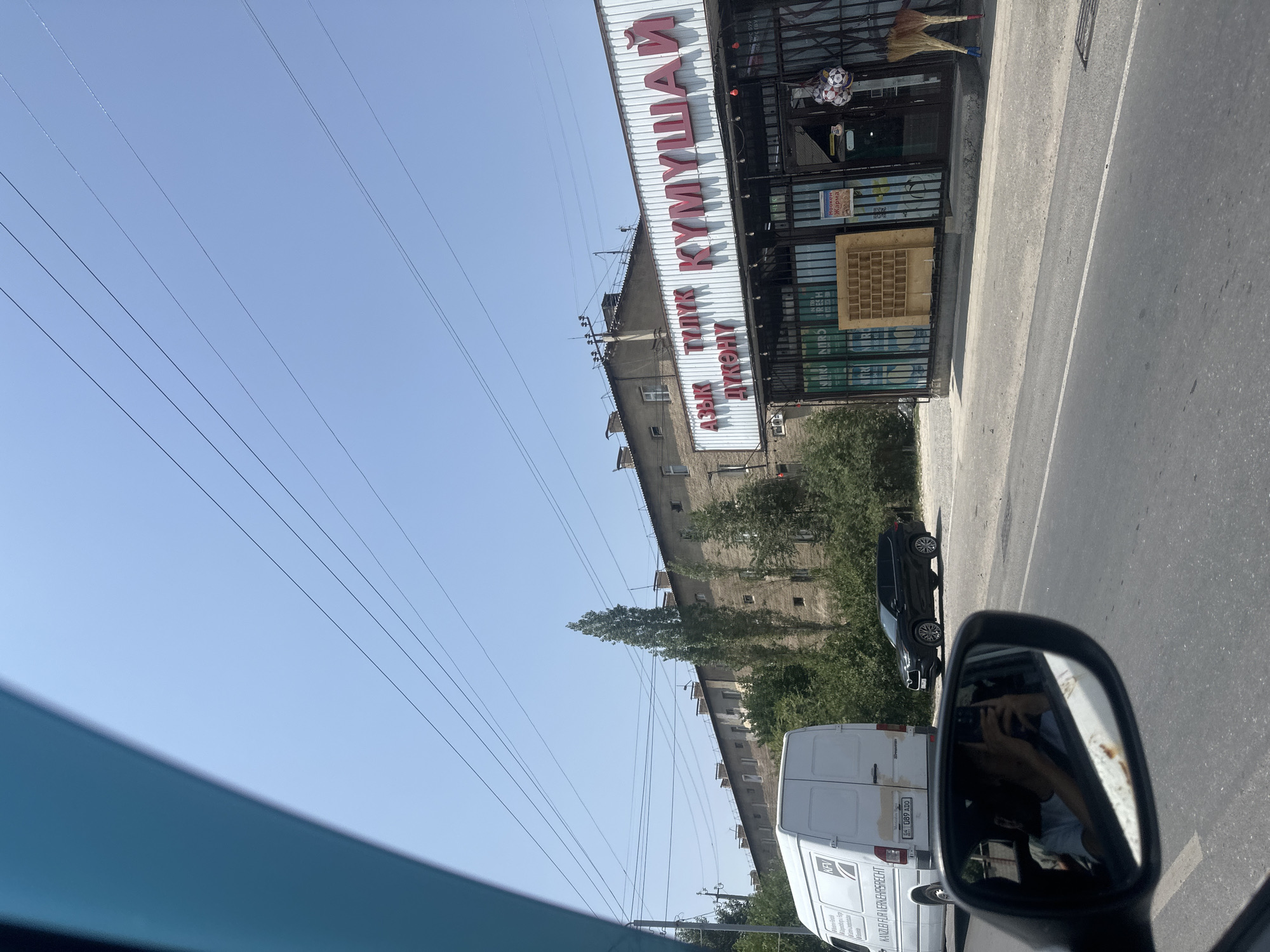 The image captures a street scene in Manas, Kyrgyzstan, under a clear blue sky. A white delivery van with "KFV KANZLER FUR VERKEHRSRECHT" written on its side is parked on the left. To its right, a black SUV is parked. In the background, a long, multi-story building with a faded facade stands behind a line of trees. A storefront with red lettering in Cyrillic, which appears to read "АЗЫК ТУУЛУК КУМУШАЙ ДУКОНУ" (Food Store Kumushai), is visible in the center right of the frame. A person is reflected in the car's side mirror, holding a phone. A broom and soccer balls are hanging outside the store. Overhead, power lines crisscross the sky.