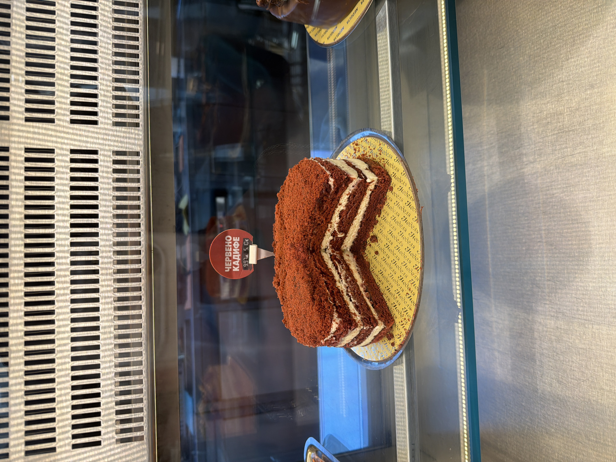 The image, presented with a 90-degree clockwise rotation, showcases a delectable slice of red velvet cake, prominently displayed within a glass refrigerated case, likely in a bakery or cafe. The cake features several layers of rich, dark red sponge alternating with generous cream cheese frosting, and its exterior is generously coated in red cake crumbs.
The cake rests on a round, yellow-patterned plate or liner, which is visibly branded with the repeated Cyrillic word "НЕДЕЛЯ" (Nedelya), translating to "Sunday" or "Week," suggesting a brand name. A small, red circular price tag is attached to the cake. The text on the tag reads "ЧЕРВЕНО КАДИФЕ" (Cherveno Kadife), which means "Red Velvet" in Bulgarian, identifying the cake type. Below the name, the price is listed as "9.97 лв." (Bulgarian Lev) and "5.10€" (Euro), indicating the cost.
The setting appears to be an indoor commercial space, with the display case featuring a metallic grated panel on the left and a reflective glass front. A vertical strip of bright LED lights illuminates the interior of the case, drawing attention to the dessert. The background, seen through the glass, shows blurred reflections of the environment, including other items or parts of the establishment. The image primarily focuses on the cake and its presentation, highlighting it for sale. The use of Bulgarian text and currency confirms the location context of Plovdiv, Bulgaria.