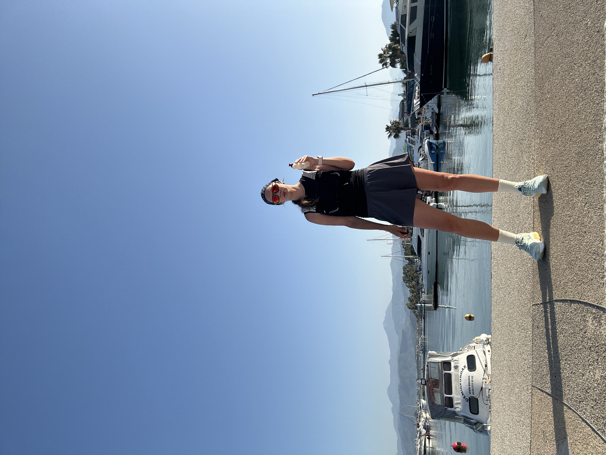 A woman in sportswear stands on a concrete pier, holding a water bottle. She wears a black top, a gray skirt, white socks, and light-colored running shoes. She also has a patterned headscarf and red sunglasses. Behind her is a marina filled with various boats and yachts, with mountains visible in the background. The location is Fethiye, Turkey, based on the appearance of the marina and the surrounding landscape. A white rescue boat with Turkish writing is docked near the pier.