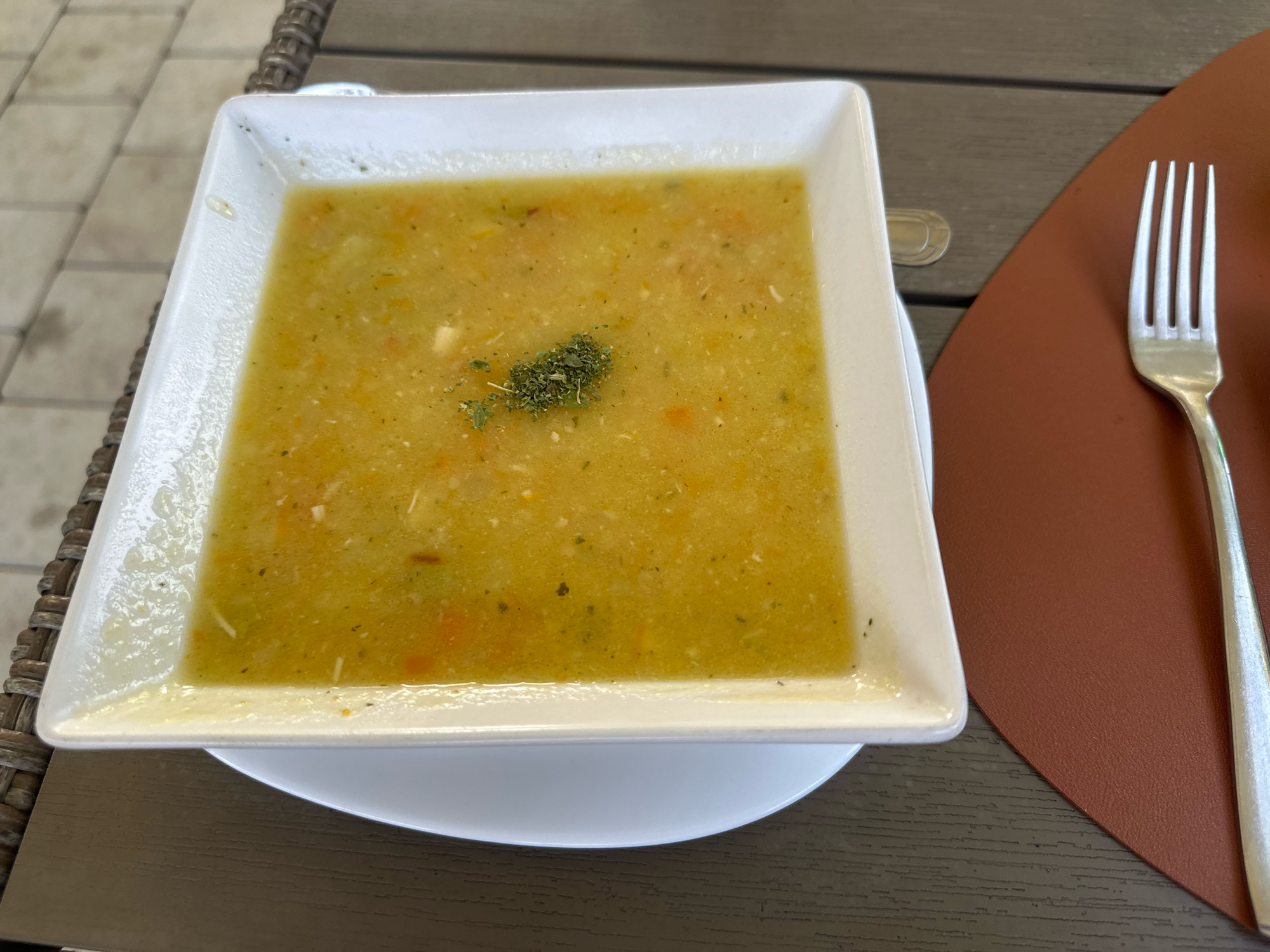 A square white bowl of yellow soup, garnished with dried herbs, sits on a white plate. The bowl and plate are on a dark brown textured table. To the right of the bowl, a silver fork rests on a brown placemat. The background shows a woven chair and a tiled walkway. The scene is set outdoors, likely at a cafe or restaurant in Topola Municipality, Serbia. There are no people or animals visible in the image.
