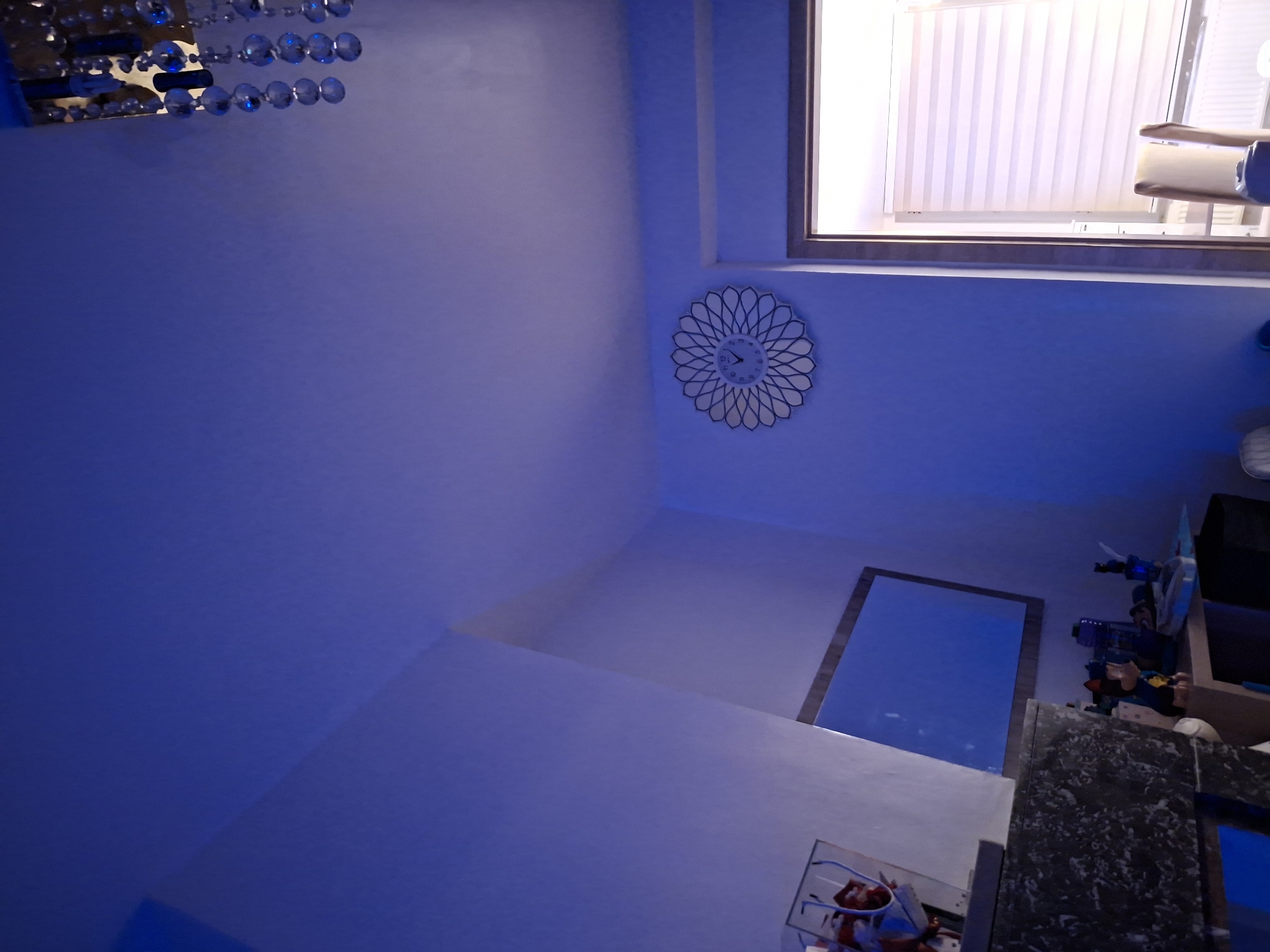 The image shows an indoor scene, likely a residential room, bathed in blue light. A ceiling with a subtle slope is visible at the top, adorned with a decorative chandelier featuring clear beads. On the right wall, a circular clock with a white, patterned face and black hands is mounted, indicating a time between 1:00 and 2:00. To the right of the clock, a doorway leads to a room with a window covered by a translucent, horizontal-striped blind. A radiator is visible beneath the window, and two chairs with light-colored upholstery are partially in view. On the left side of the room, a mirror with a dark frame hangs on the wall. Below the mirror, a dark, patterned surface, possibly a dresser or shelf, is present. On this surface, a photo frame is visible, containing what appear to be people. Further to the right and lower, a shelf or desk holds various objects, including figurines, a blue cup, and other items that are not clearly identifiable due to the lighting. The overall impression is of a room decorated with a modern aesthetic, potentially a child's room given the figurines. The location is Evere, Belgium.
