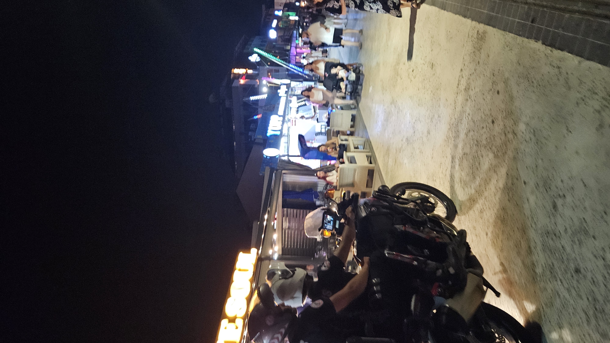 At night on a street in Fethiye, Turkey, two individuals wearing police uniforms are on a motorcycle. The street is lined with businesses, including a restaurant or bar with outdoor seating and illuminated signage that reads "TES SOUL". Further down the street, other businesses with neon lights are visible. Several pedestrians are walking along the street. One group includes a woman pushing a stroller with a child. Another group consists of people seated at tables outside a venue. The ground is a light-colored paved surface, and the overall lighting suggests an urban evening setting.