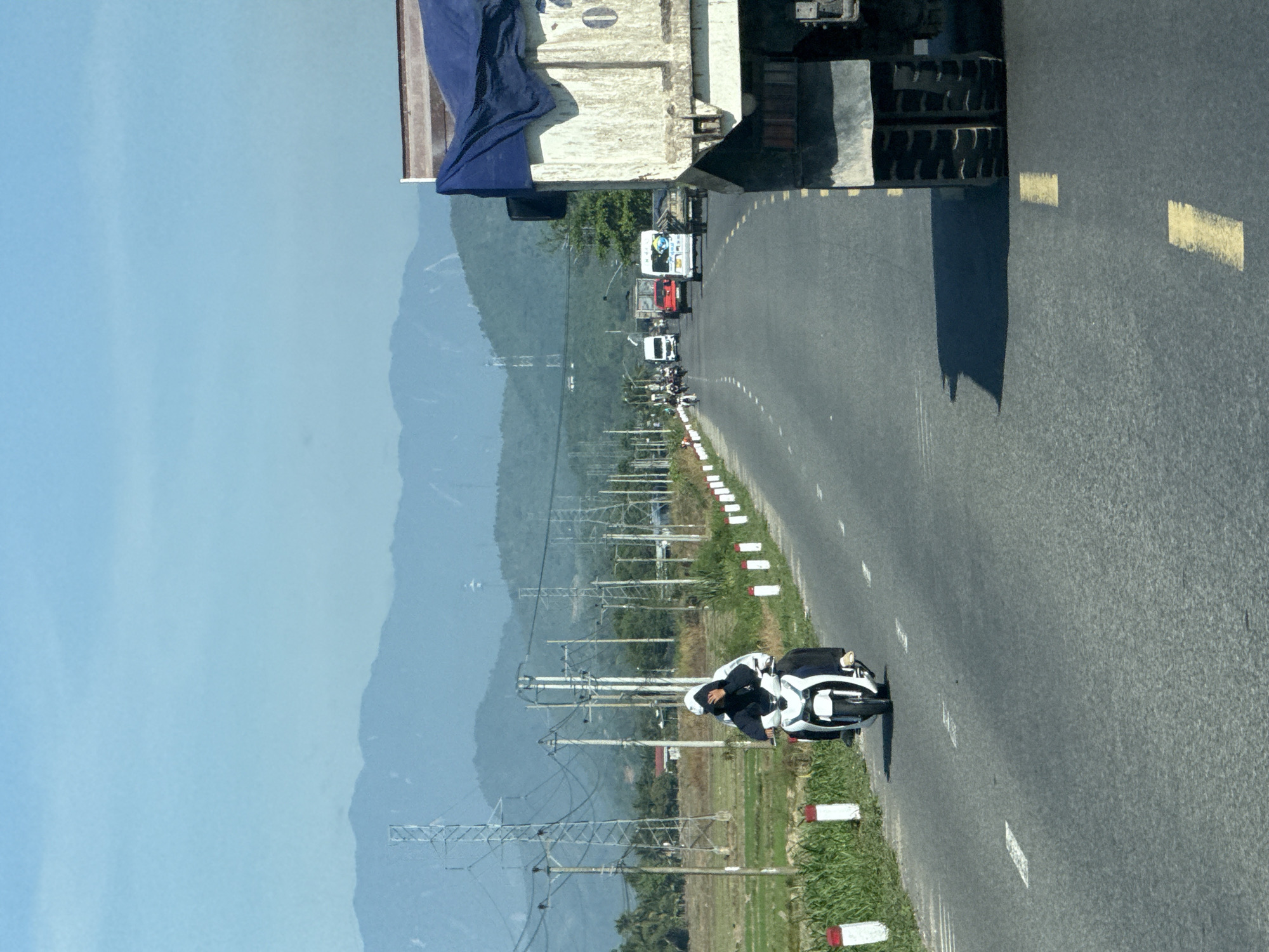 In the city of Dien Lac, Vietnam, on a paved road, a scooter carrying two individuals travels towards the camera. The person in front wears a black and white jacket and a black head covering. The person behind them is covered in a black jacket and a white head covering. A large truck, partially visible on the right side of the image, is moving away from the camera. The road has a white dashed line and a yellow dashed line marking the lanes. In the background, there are lush green fields, tall electrical towers, and distant mountains under a clear blue sky. Other vehicles, including cars and motorcycles, are visible on the road in the distance.